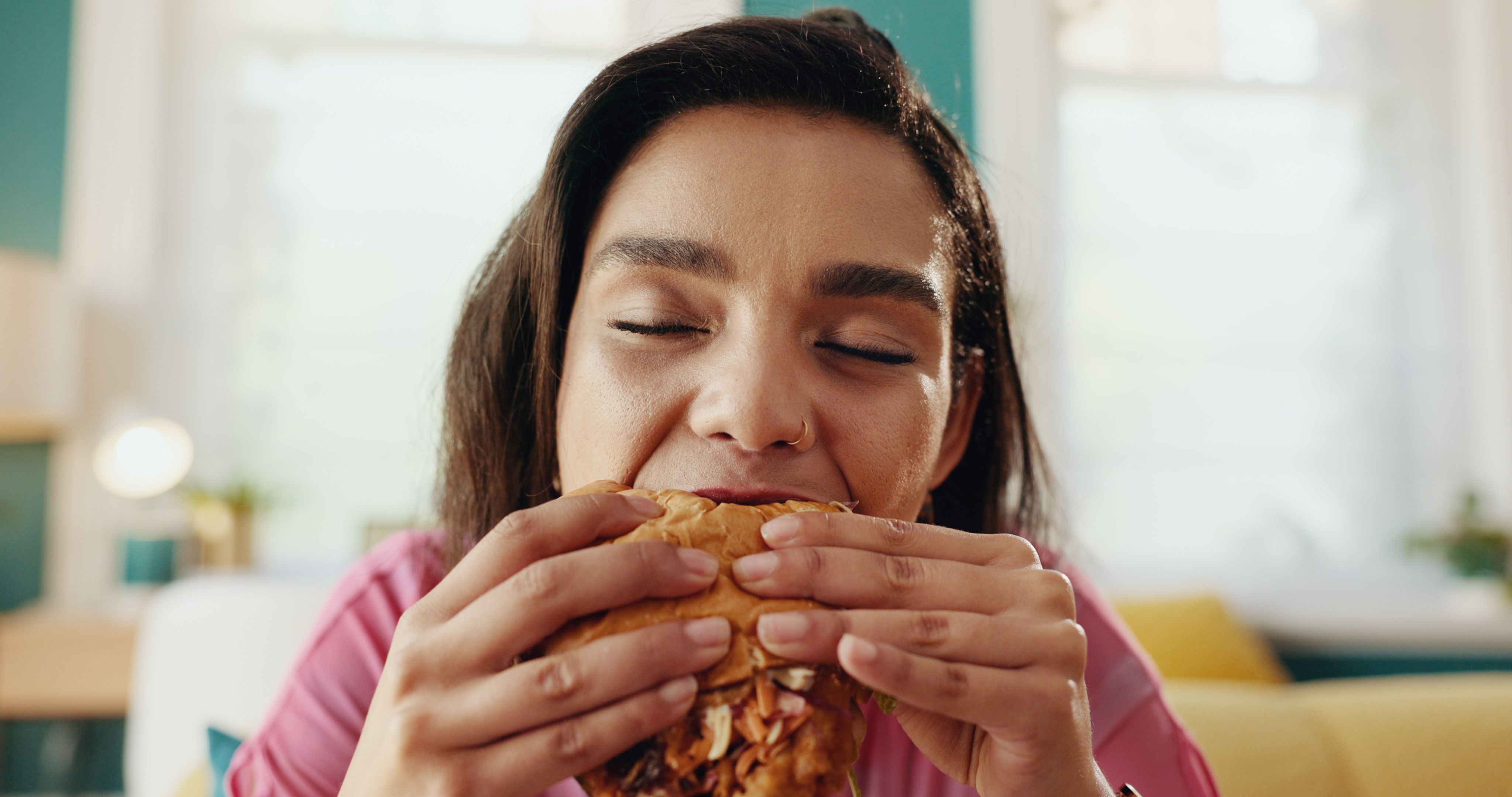 Home, burger and hungry woman eating fast food, unhealthy meal and takeaway to relax in living room. Nutrition, hamburger and girl enjoy lunch order, beef sandwich or meat for diet or snack addiction