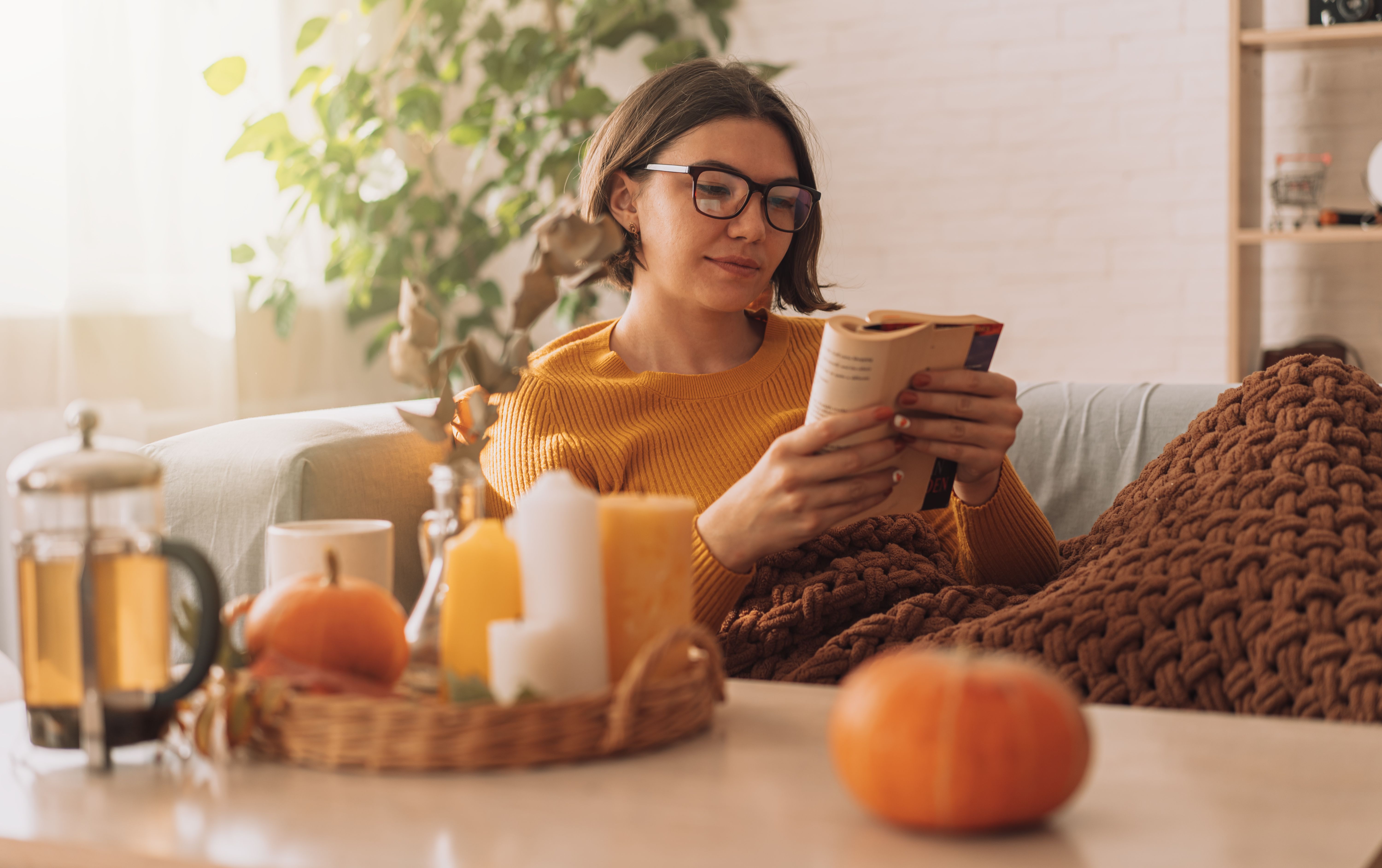 Lady reads a book in blanket on the sofa in front of teapot and candles