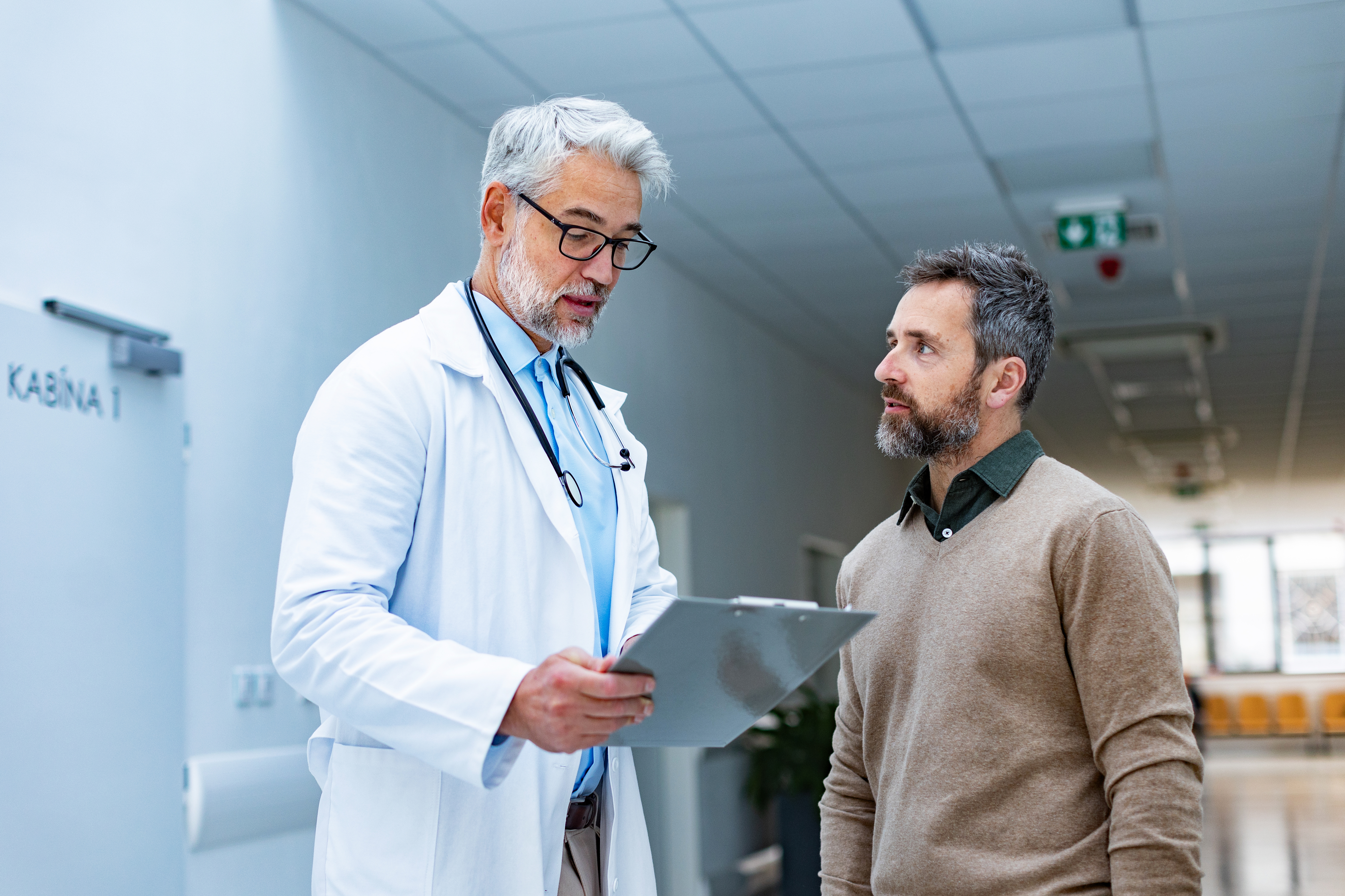 Doctor talking with patient in hospital corridor. Handsome doctor with gray hair holding clipboard with test result, consulting with man, standing in modern clinic.