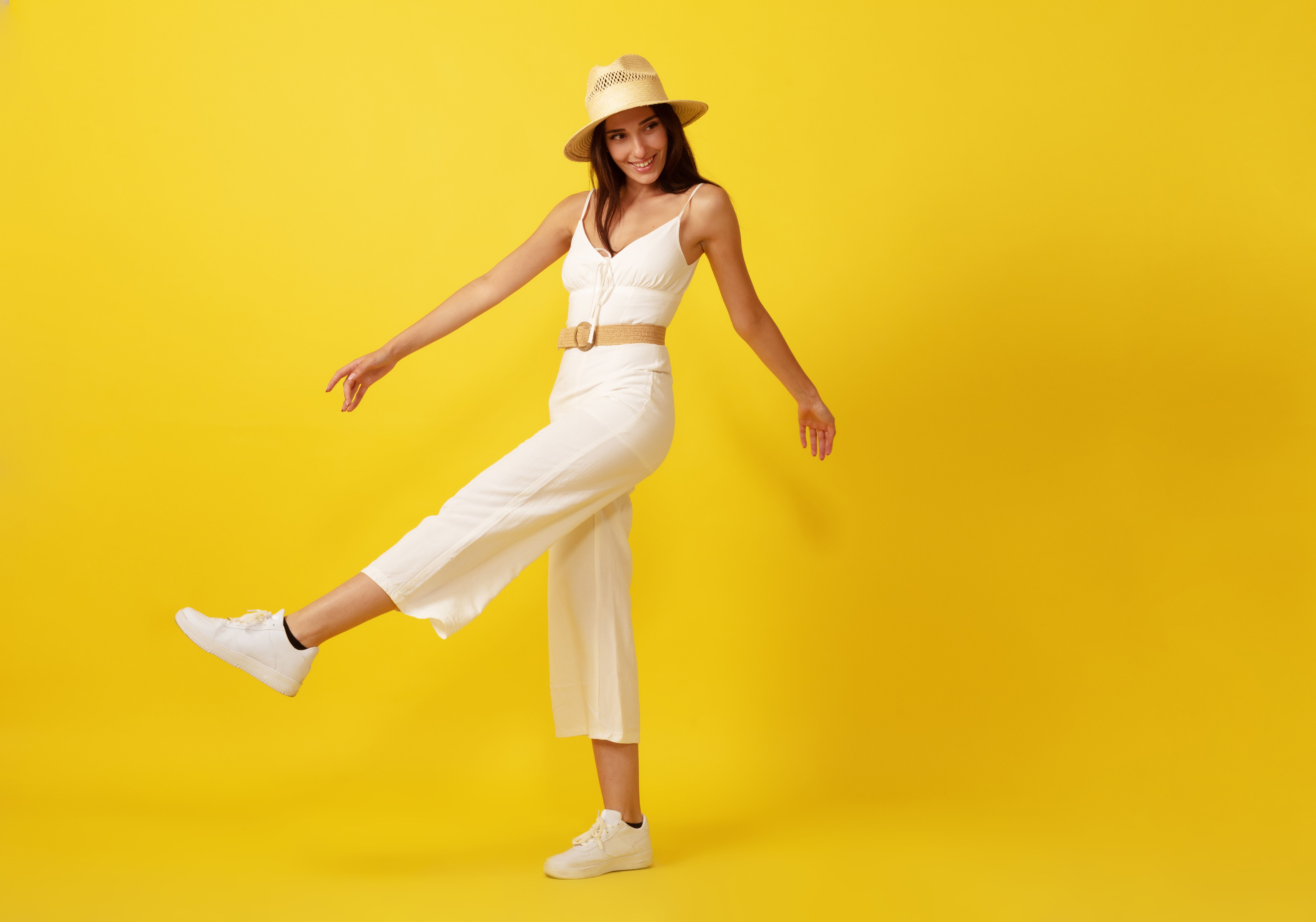 Beautiful young woman walking with straw hat in white dress front of yellow background.