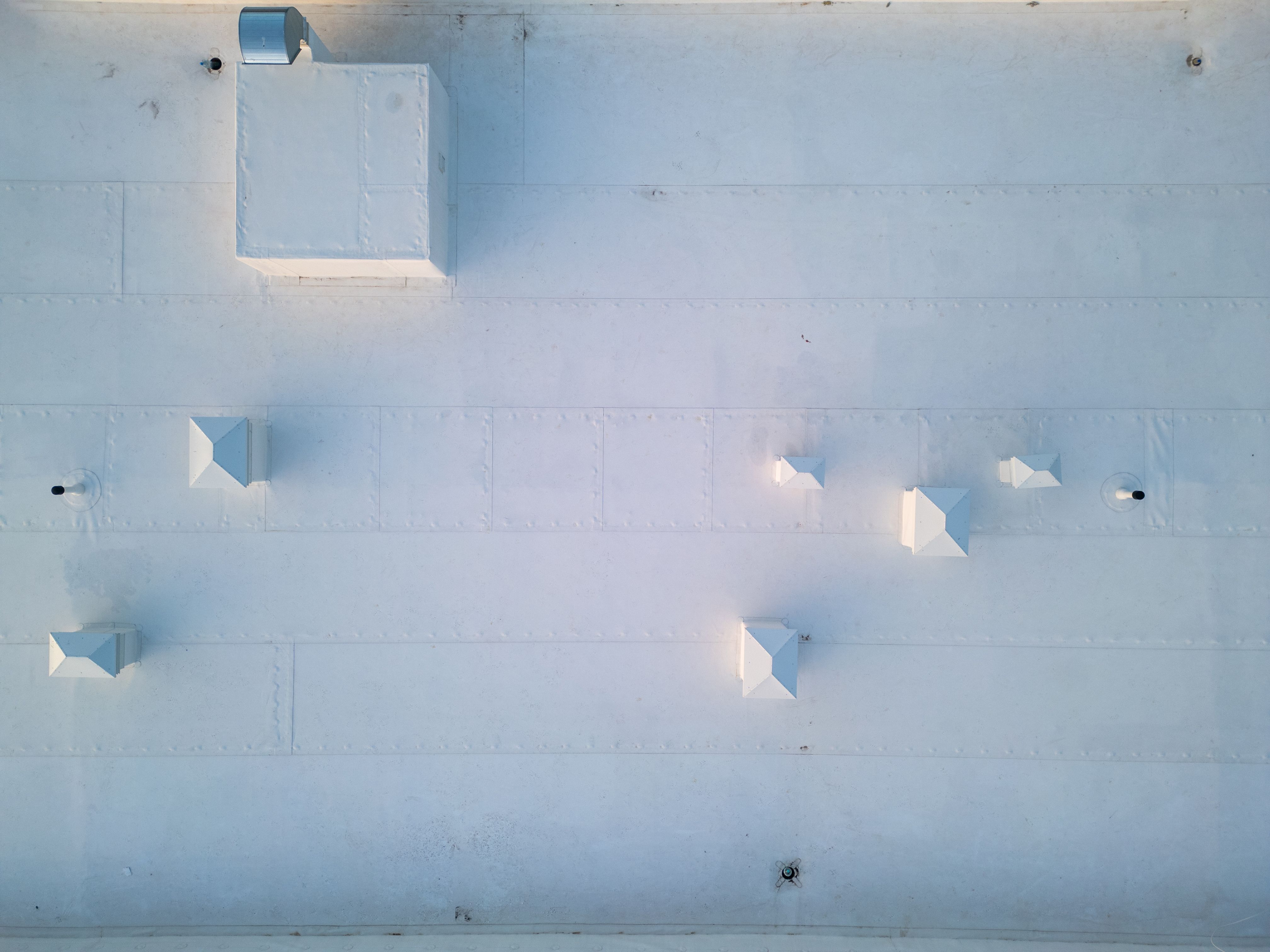Aerial view of a white industrial rooftop showcasing a clean, minimalistic design with multiple vents and structures. The symmetrical layout and geometric patterns create a modern architectural aesthetic, captured from a drone perspective