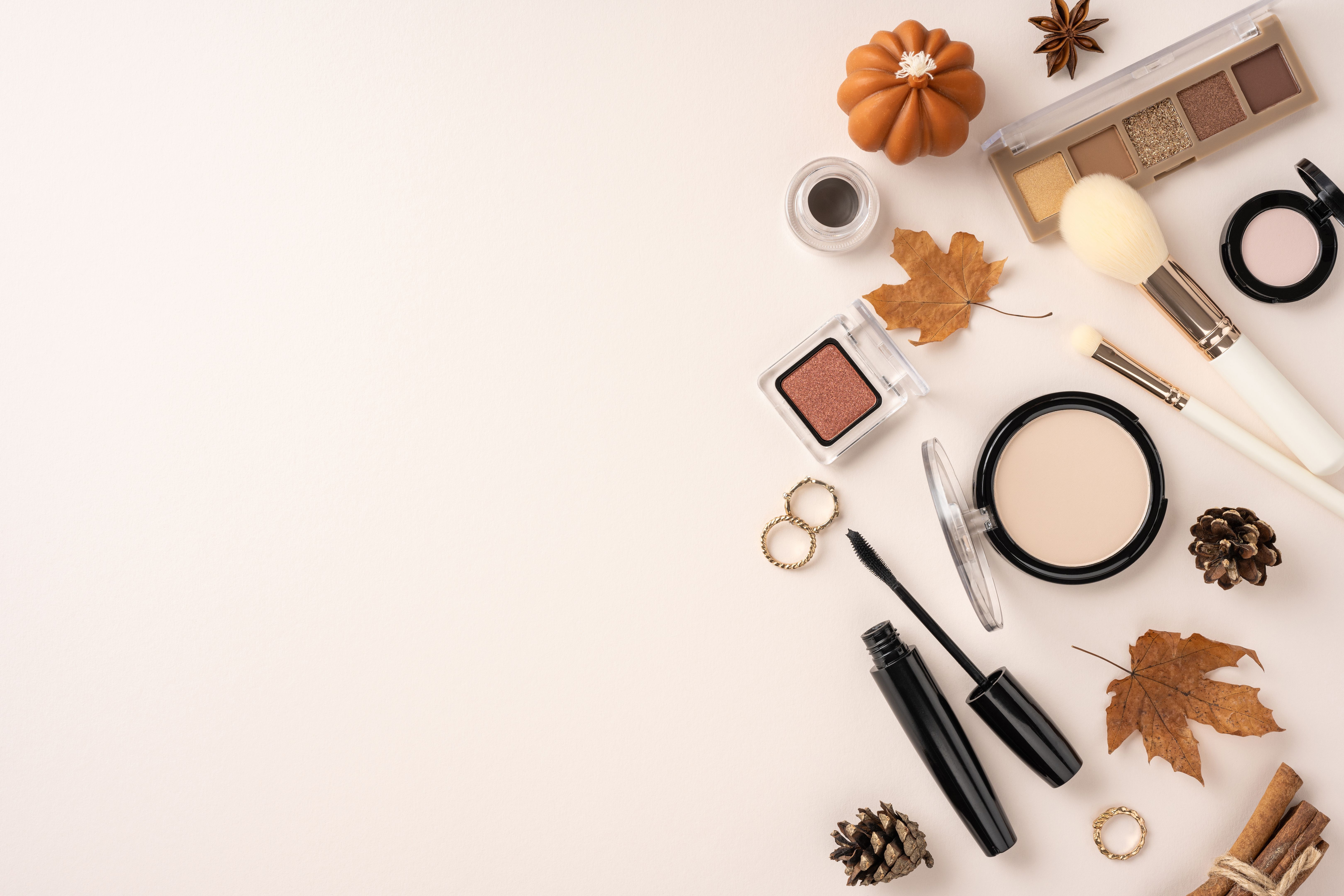 Flat lay of various autumn-themed makeup products and accessories on a beige background, featuring leaves, pine cones, and cosmetics
