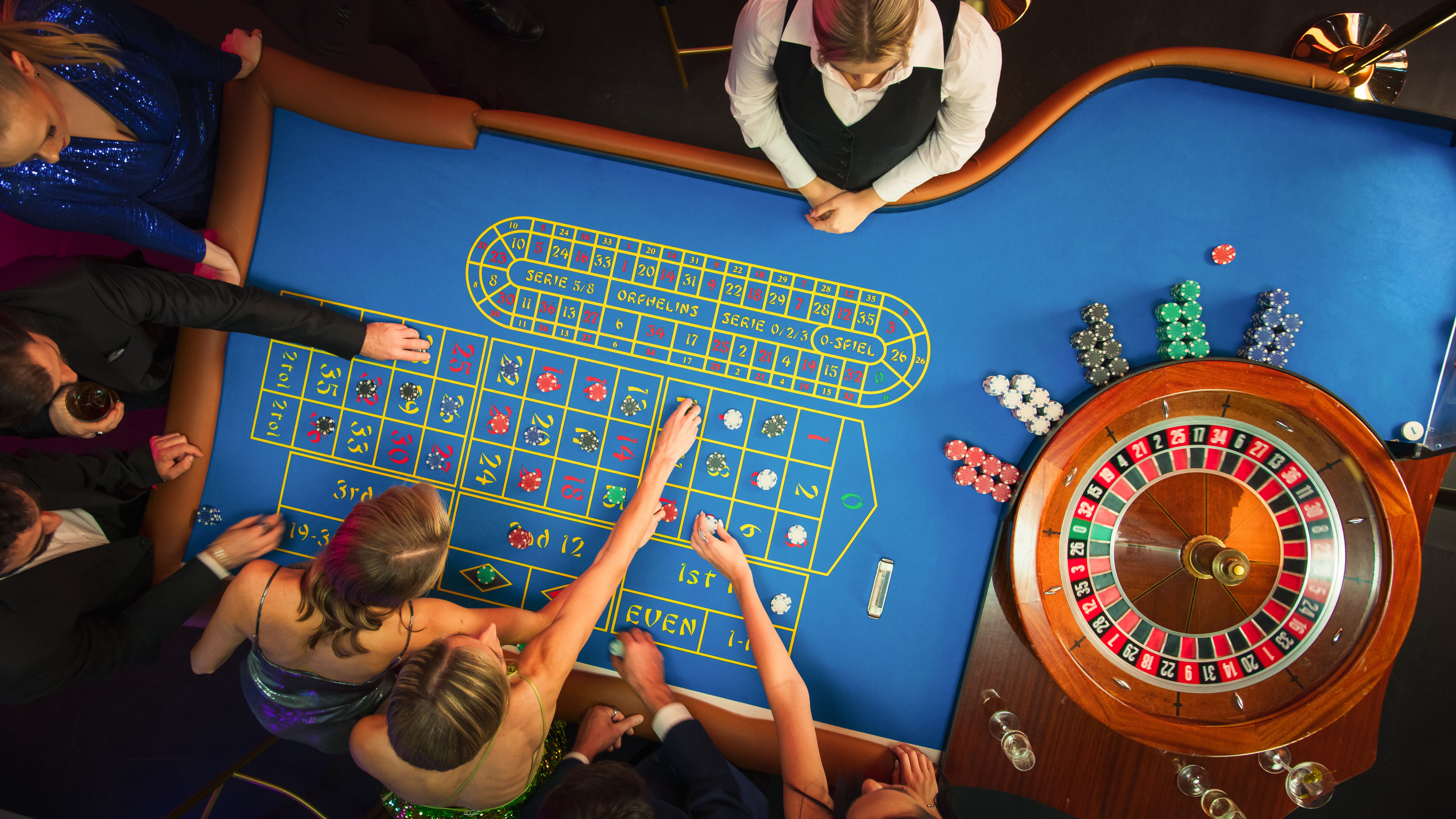 Top Down View: Luxurious Casino Male and Female Guests Putting Betting Chips on a Gambling Table, Trying to Predict the Outcome of a Roulette Game Top Down View: Luxurious Casino Male and Female Guests Putting Betting Chips on a Gambling Table, Trying to Predict the Outcome of a Roulette Game
