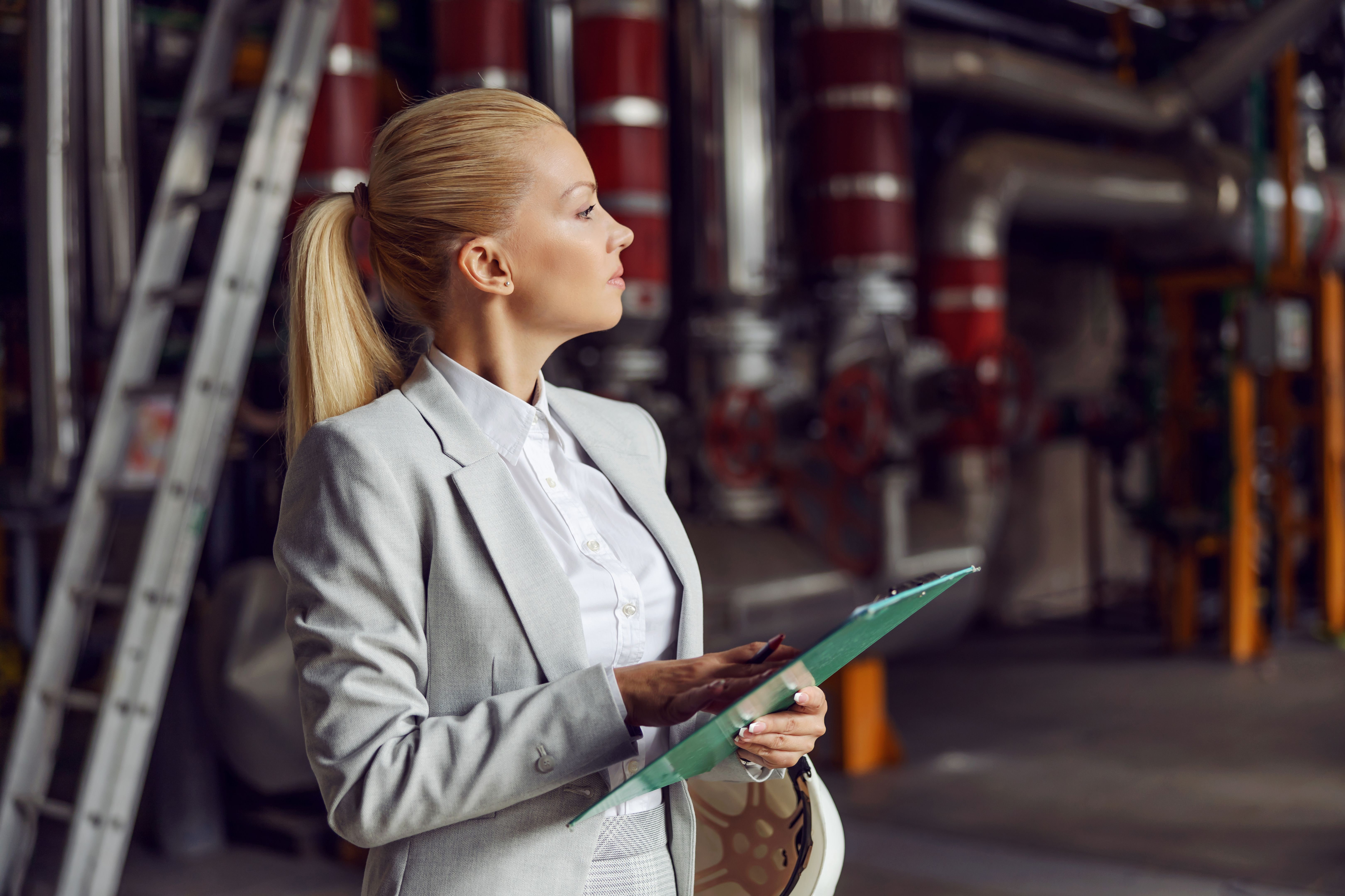 Blond businesswoman standing in heating plant, holding clipboard and checking on machinery. Blond businesswoman standing in heating plant, holding clipboard and checking on machinery.