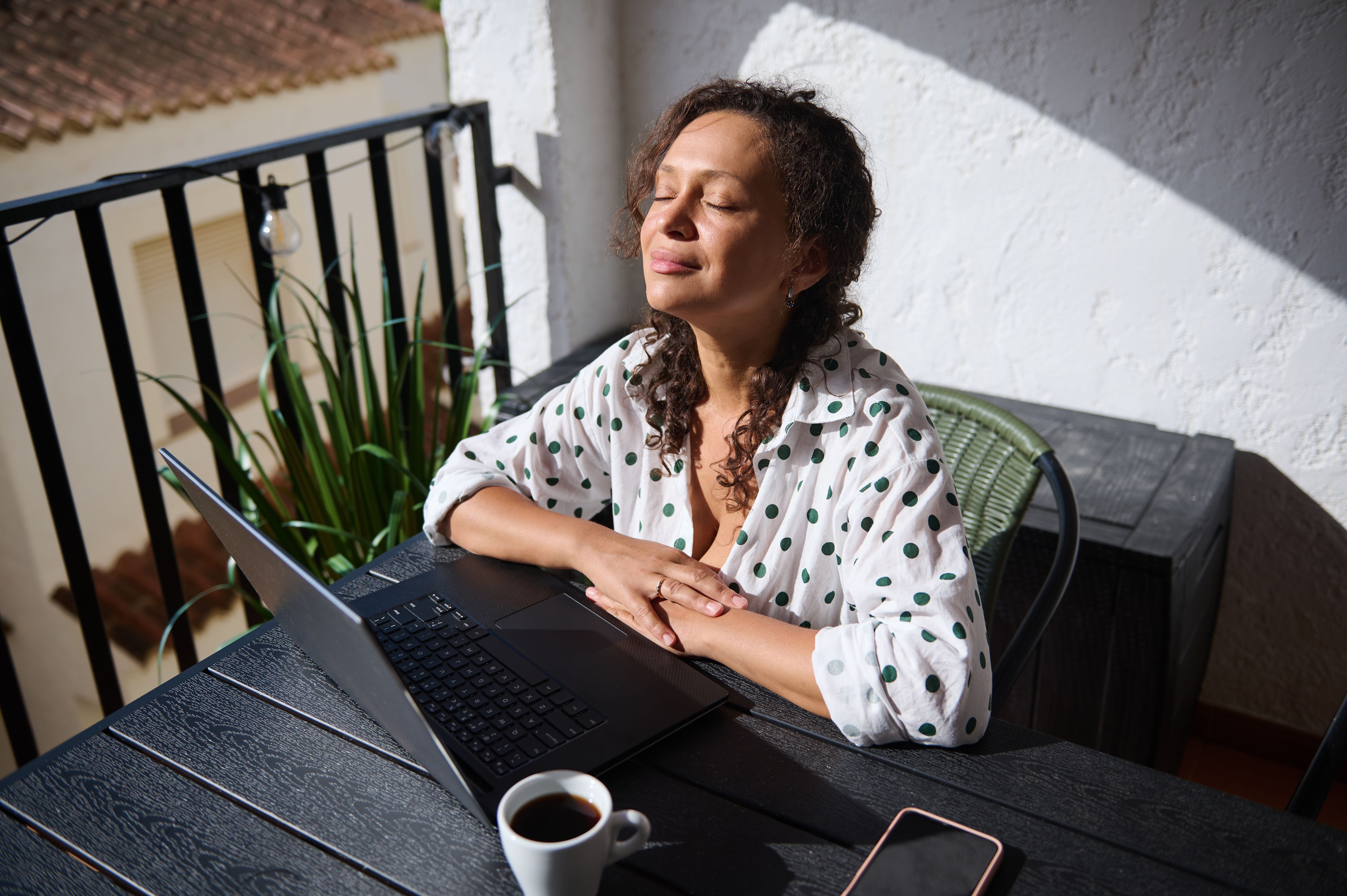 Woman enjoying a peaceful moment on balcony with laptop and coffee, absorbing the sunlight, wearing a polka dot shirt