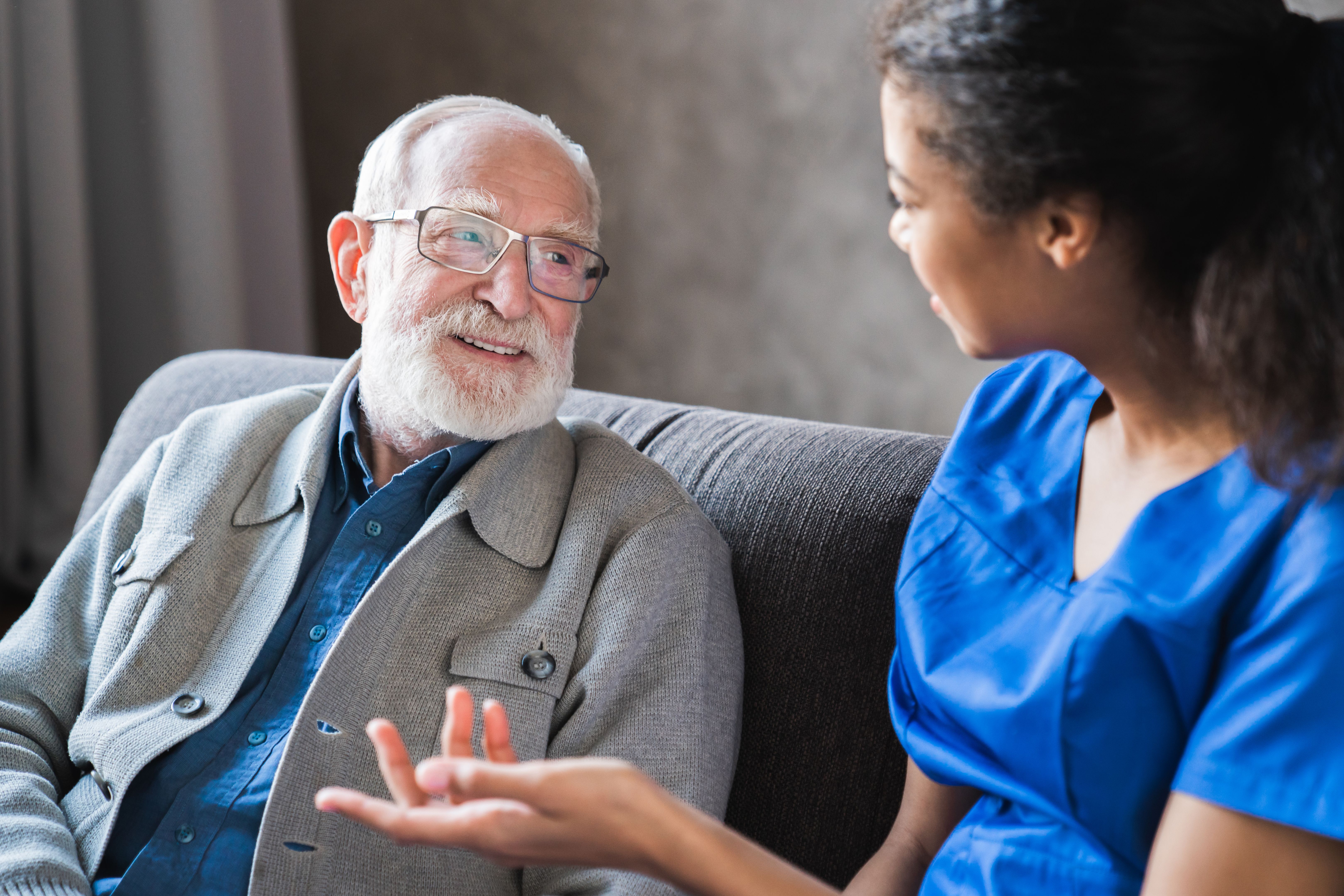 Female african professional doctor consulting senior patient grandfather during medical care visit. Young woman physician and old man talking providing medical assistance sitting on sofa. Elderly people home care concept Female african professional doctor consulting senior patient grandfather during medical care visit. Young woman physician and old man talking providing medical assistance sitting on sofa. Elderly people home care concept
