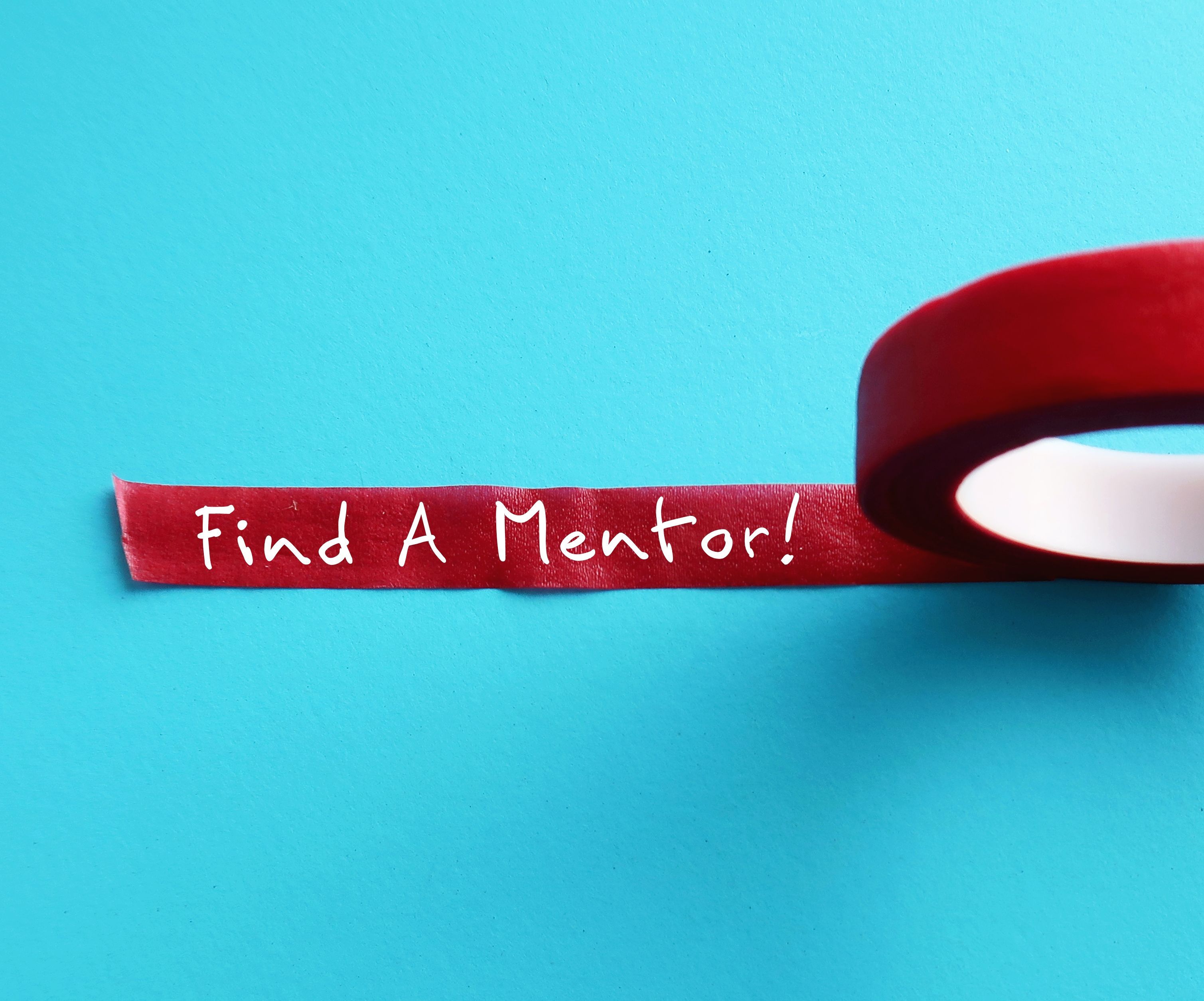 Red tape on background with handwritten text FIND A MENTOR, means a professional who informally guides a less experienced person or a mentee, to help with job requirements and long-term career goals