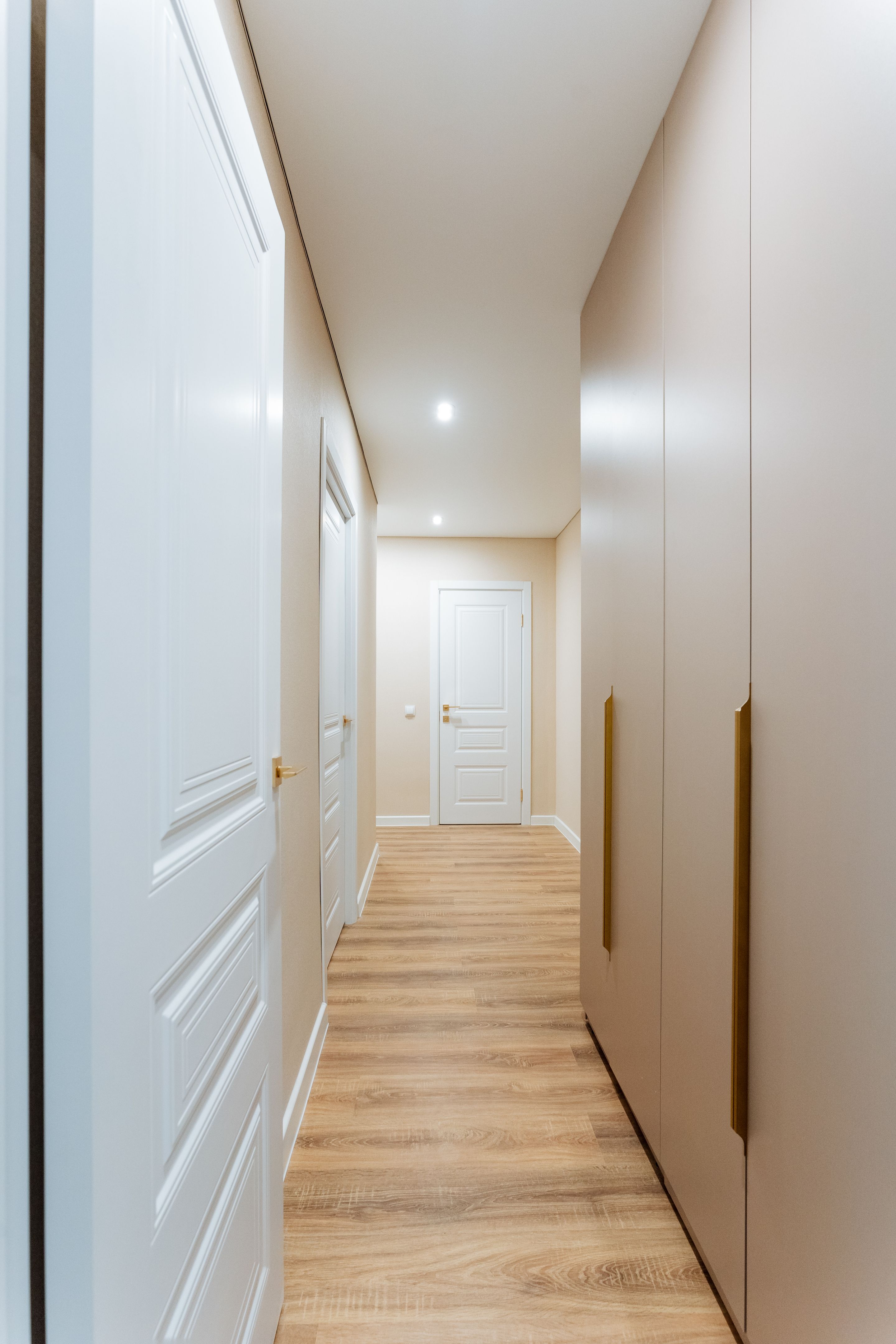 In this expansive and visually appealing long hallway, one can observe the beautiful wooden floors that add warmth and character, complemented by a series of pristine white doors
