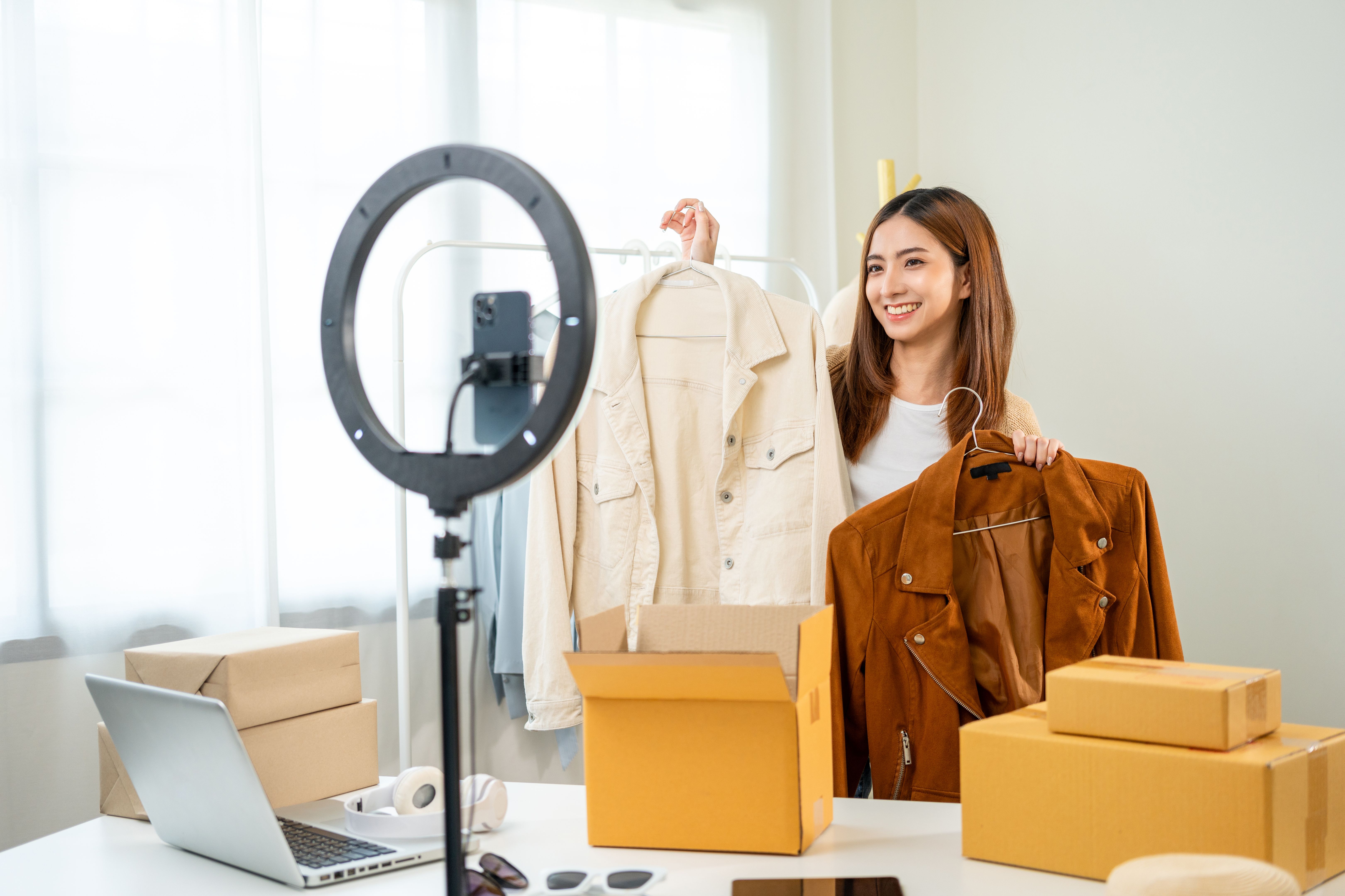 Young asian woman business owner at fashion store using smartphone live streaming for sale fashion clothing. Influencers record video review clothes for sell on social media. Online shopping concept.