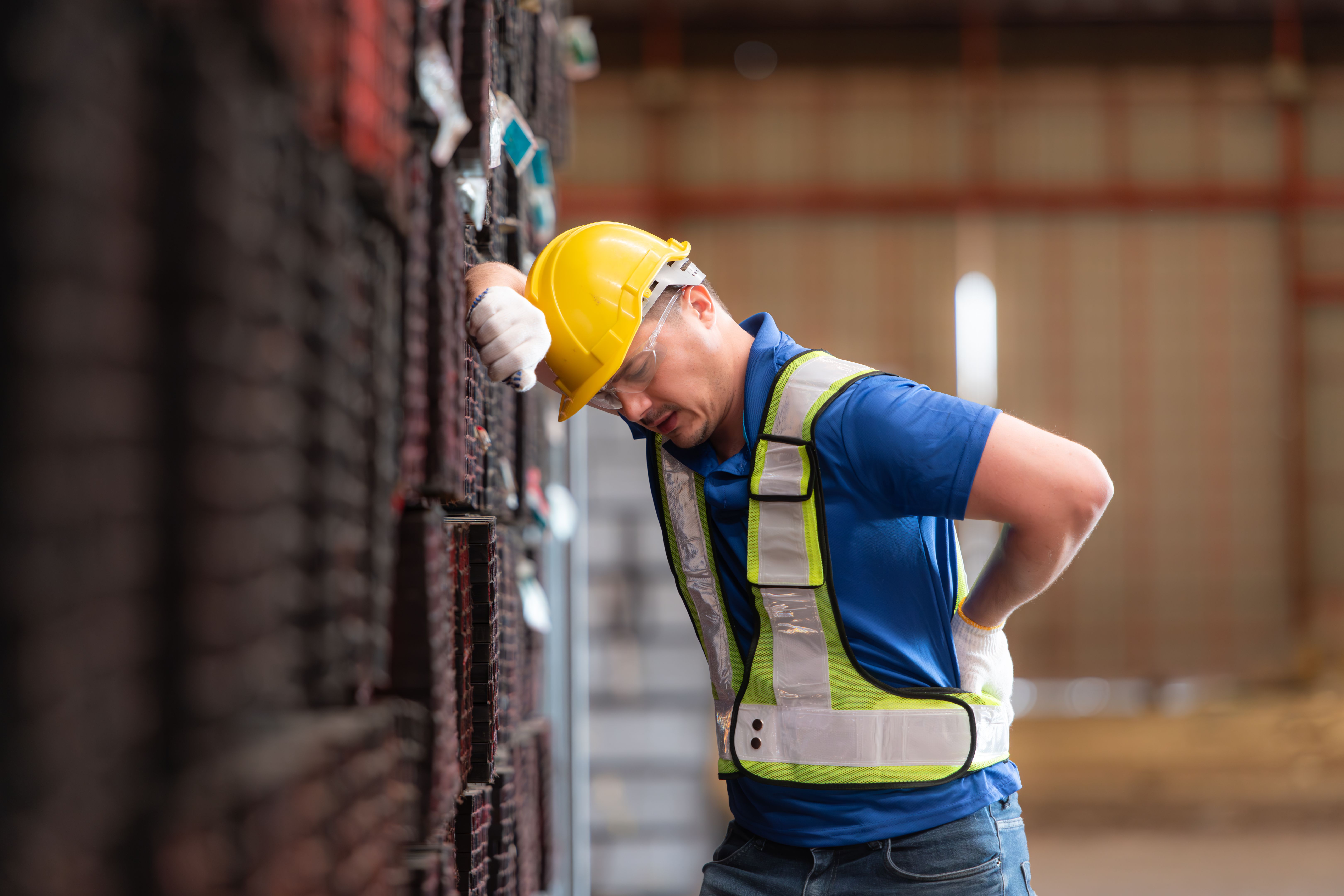Portrait of a male worker wearing a safety vest and helmet standing on a steels pallet due to back pain from working in a factory lifting heavy things.