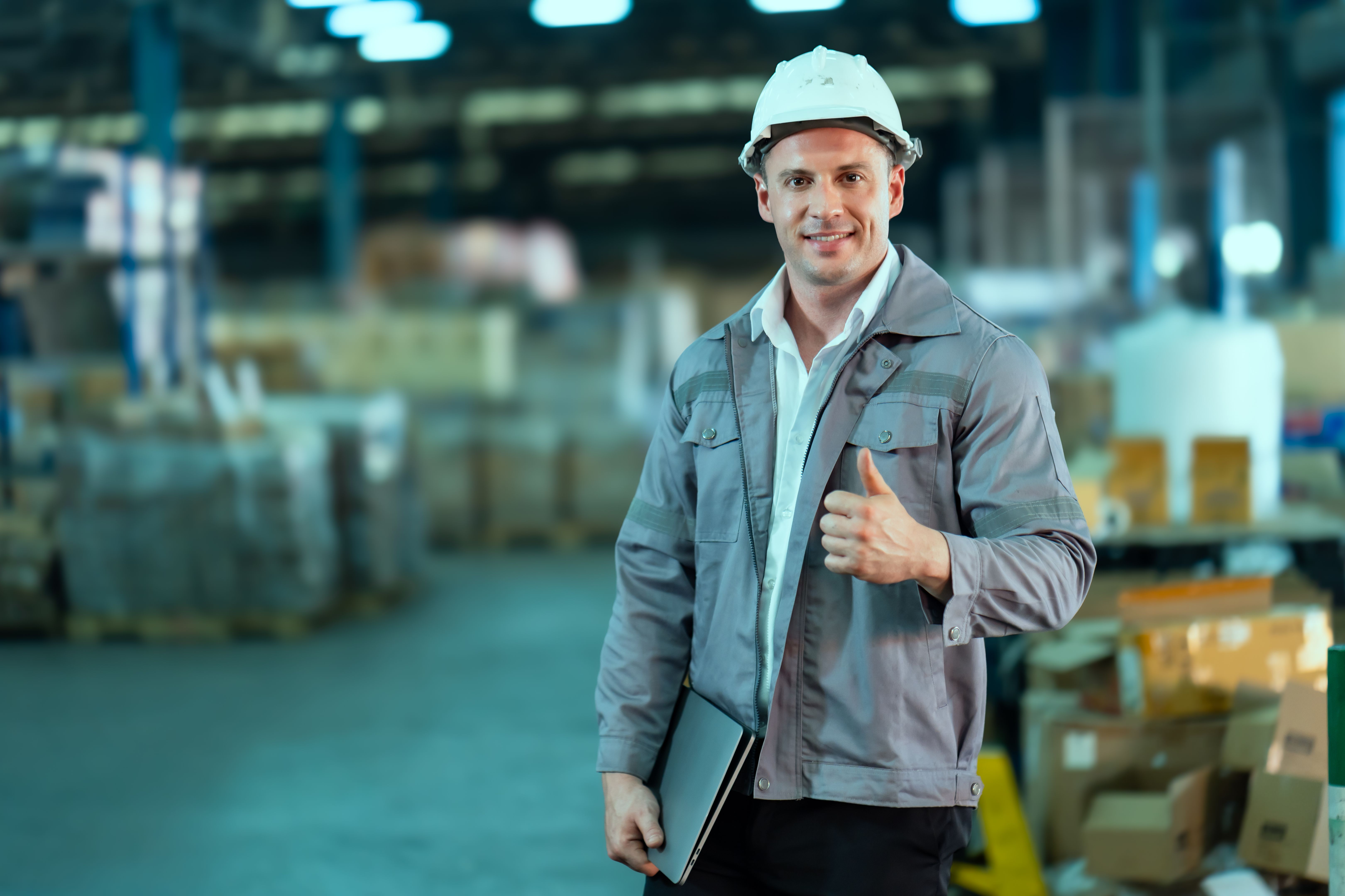 Warehouse male worker wearing helmet smiling holding laptop giving thumbs up representing confidence success professionalism teamwork logistics operations management industrial workplace business