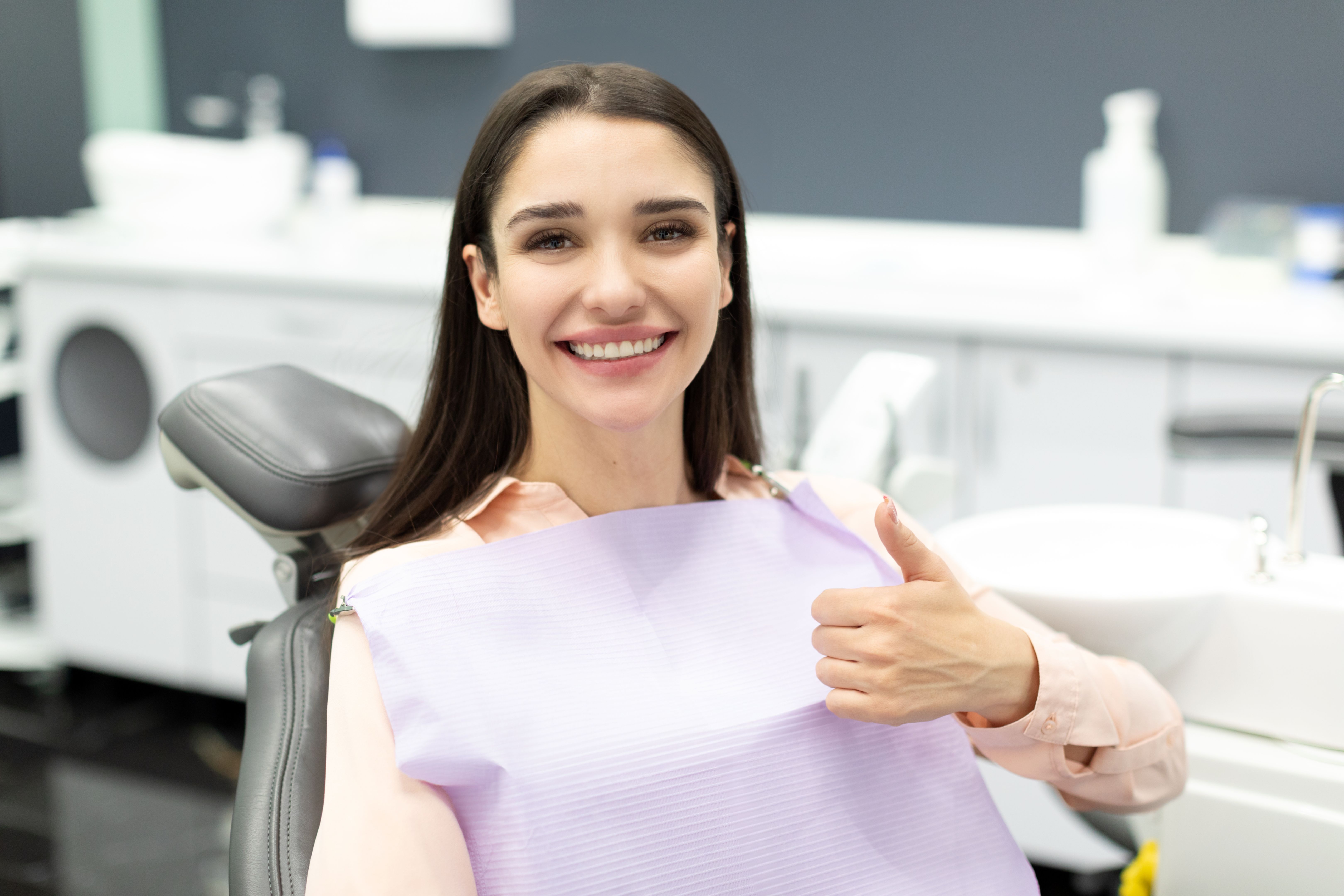 Hermosa paciente femenina feliz que muestra el pulgar hacia arriba después del tratamiento dental, sentada en la silla en la clínica estomatológica moderna después del chequeo