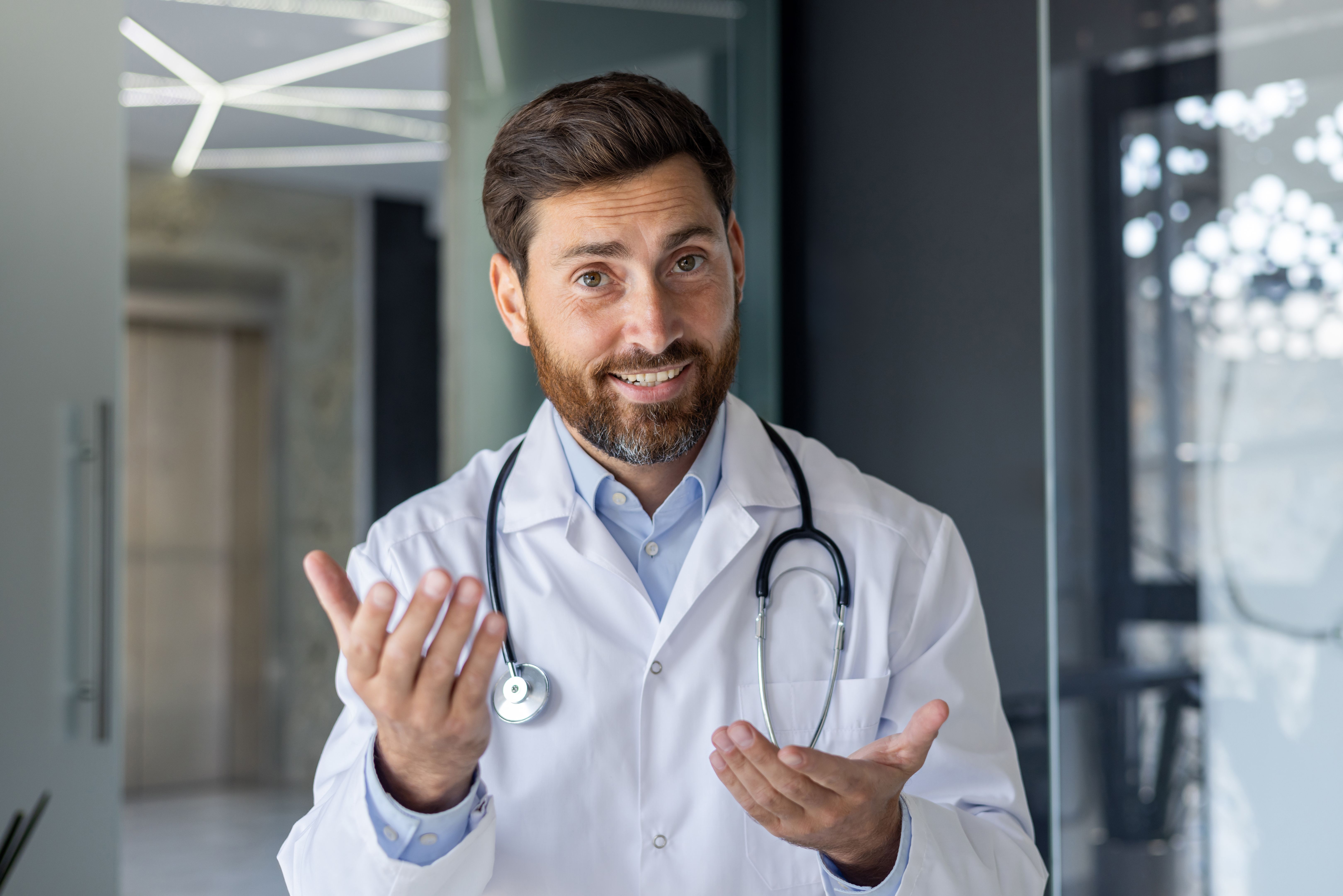 Close-up portrait of a young smiling male doctor who is in the hospital and talking via video link, conducting an online consultation and meeting, gesturing with his hands