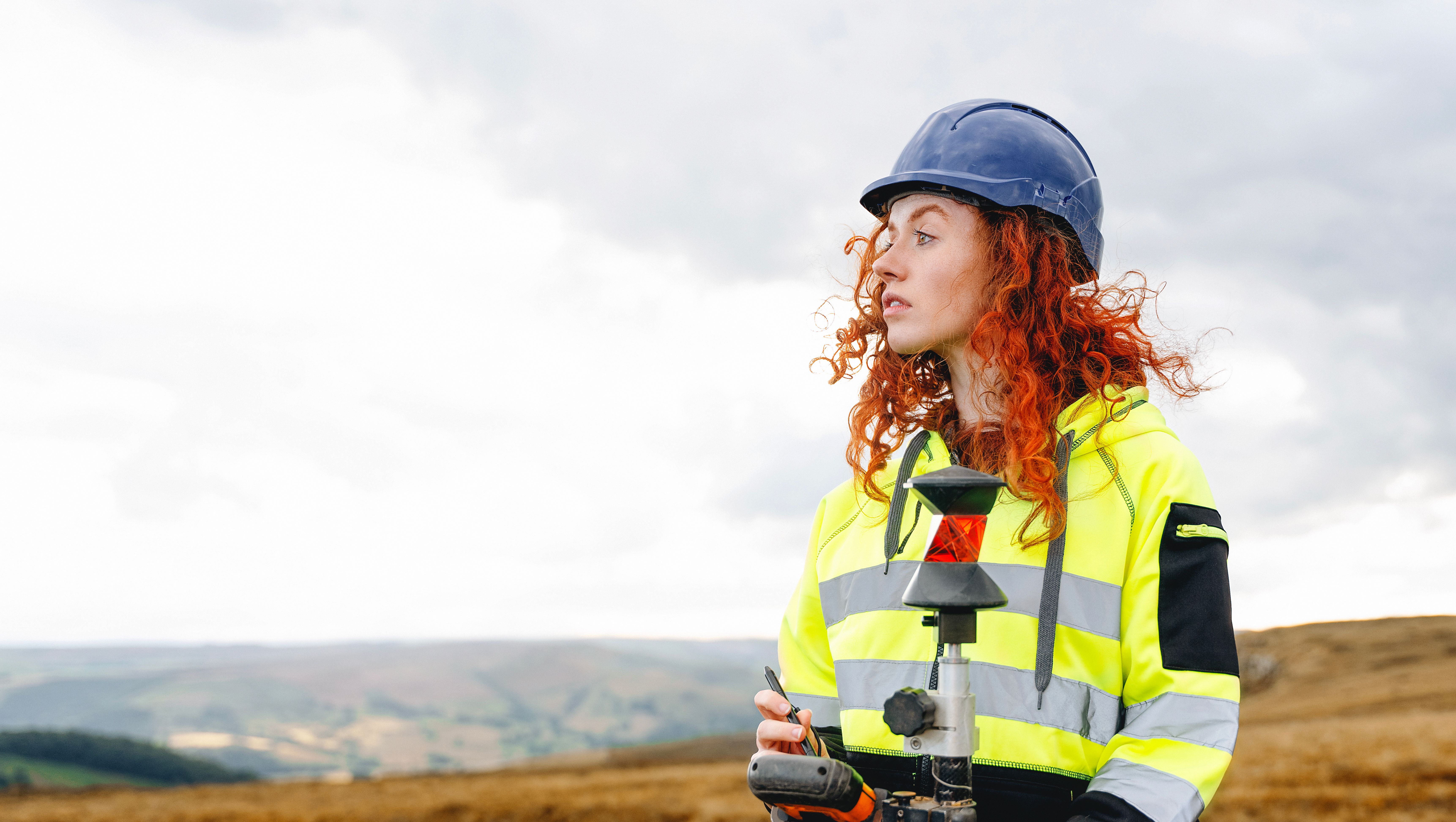 Portrait of confident female land surveyor with long hair in bright safety gear using surveying instrument to perform land survey on building site Portrait of confident female land surveyor with long hair in bright safety gear using surveying instrument to perform land survey on building site