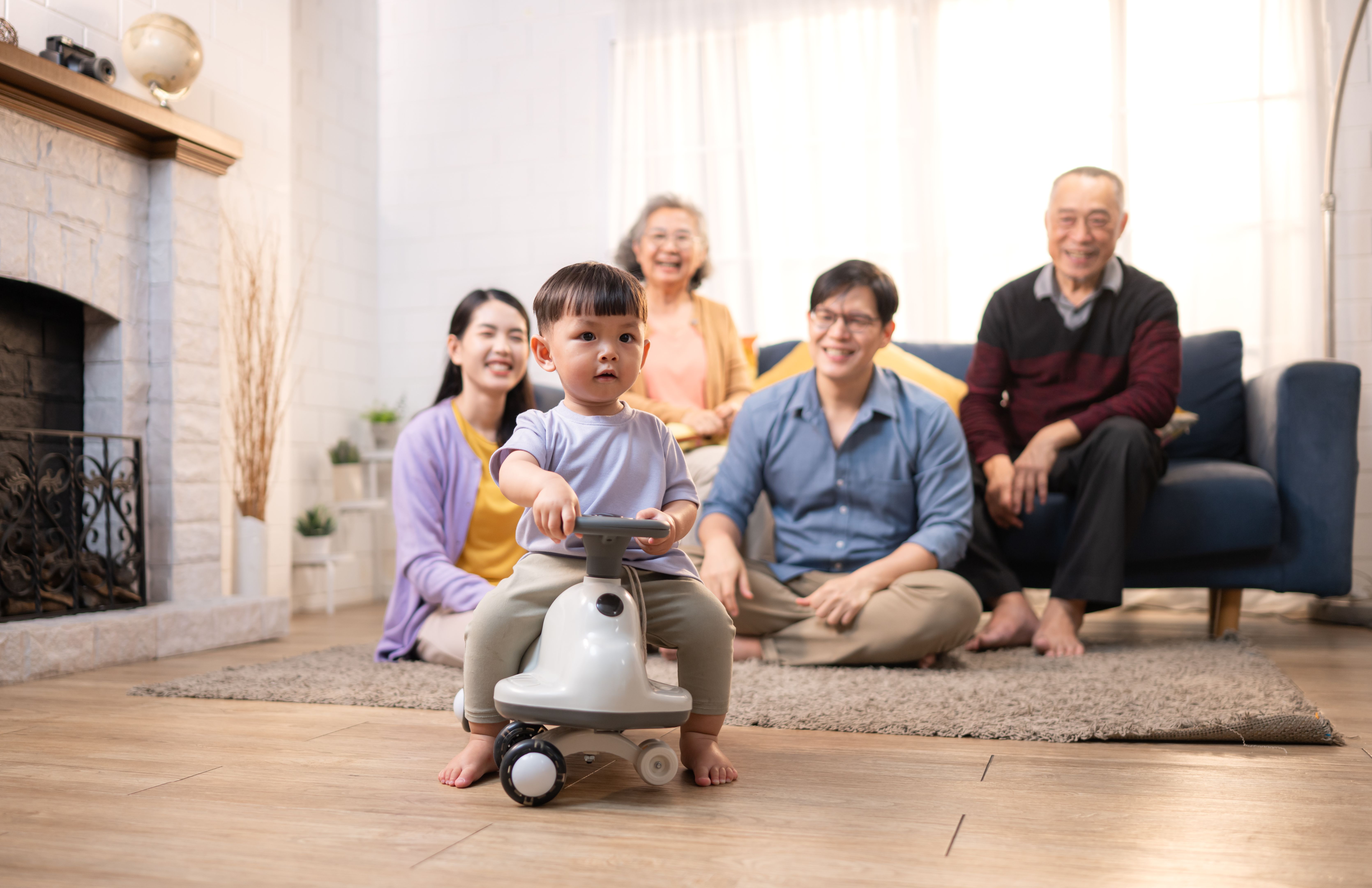 Happy multi-generational Asian family enjoying quality time together at home, cheering and smiling with a toddler riding a toy car in a cozy living room Happy multi-generational Asian family enjoying quality time together at home, cheering and smiling with a toddler riding a toy car in a cozy living room
