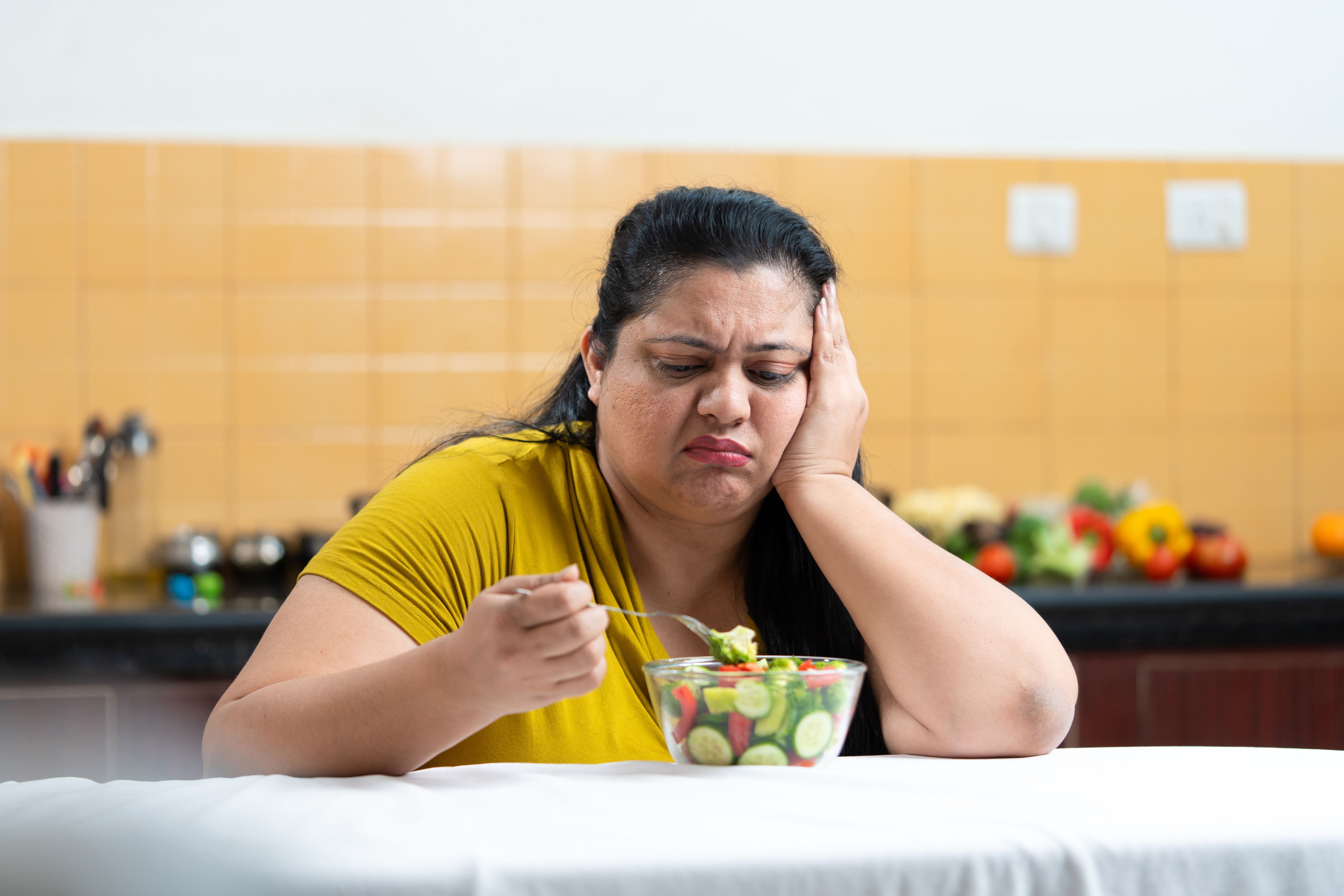 Unhappy overweight indian woman eating food salad in kitchen. Plus size female. Weight loss, Fitness and healthy diet. Unhappy overweight indian woman eating food salad in kitchen. Plus size female. Weight loss, Fitness and healthy diet.