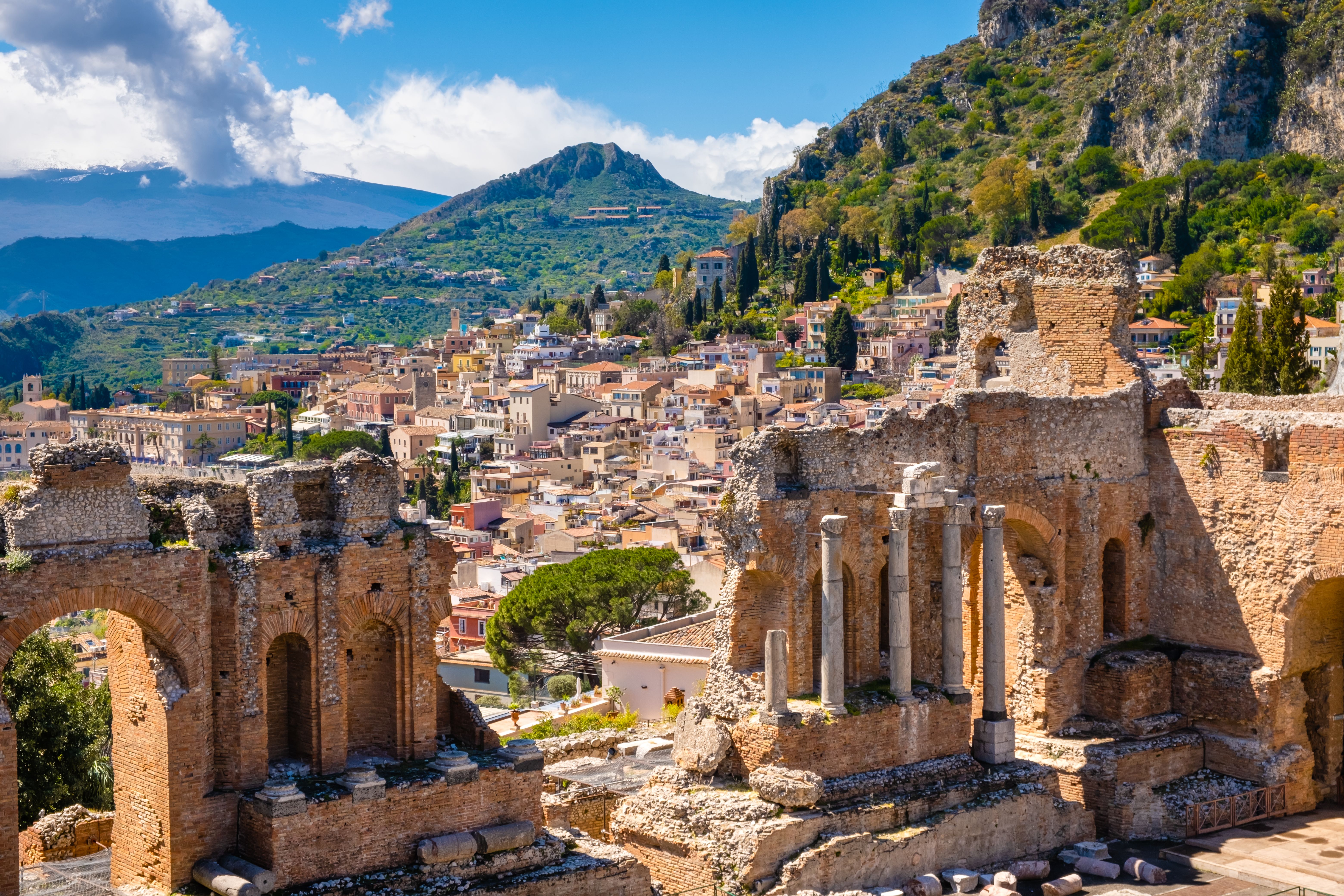 Taormina on Sicily, Italy. Ruins of ancient Greek theater, mount Etna covered with clouds. Taormina old town and mountains in background. Popular touristic destination on Sicily Taormina on Sicily, Italy. Ruins of ancient Greek theater, mount Etna covered with clouds. Taormina old town and mountains in background. Popular touristic destination on Sicily
