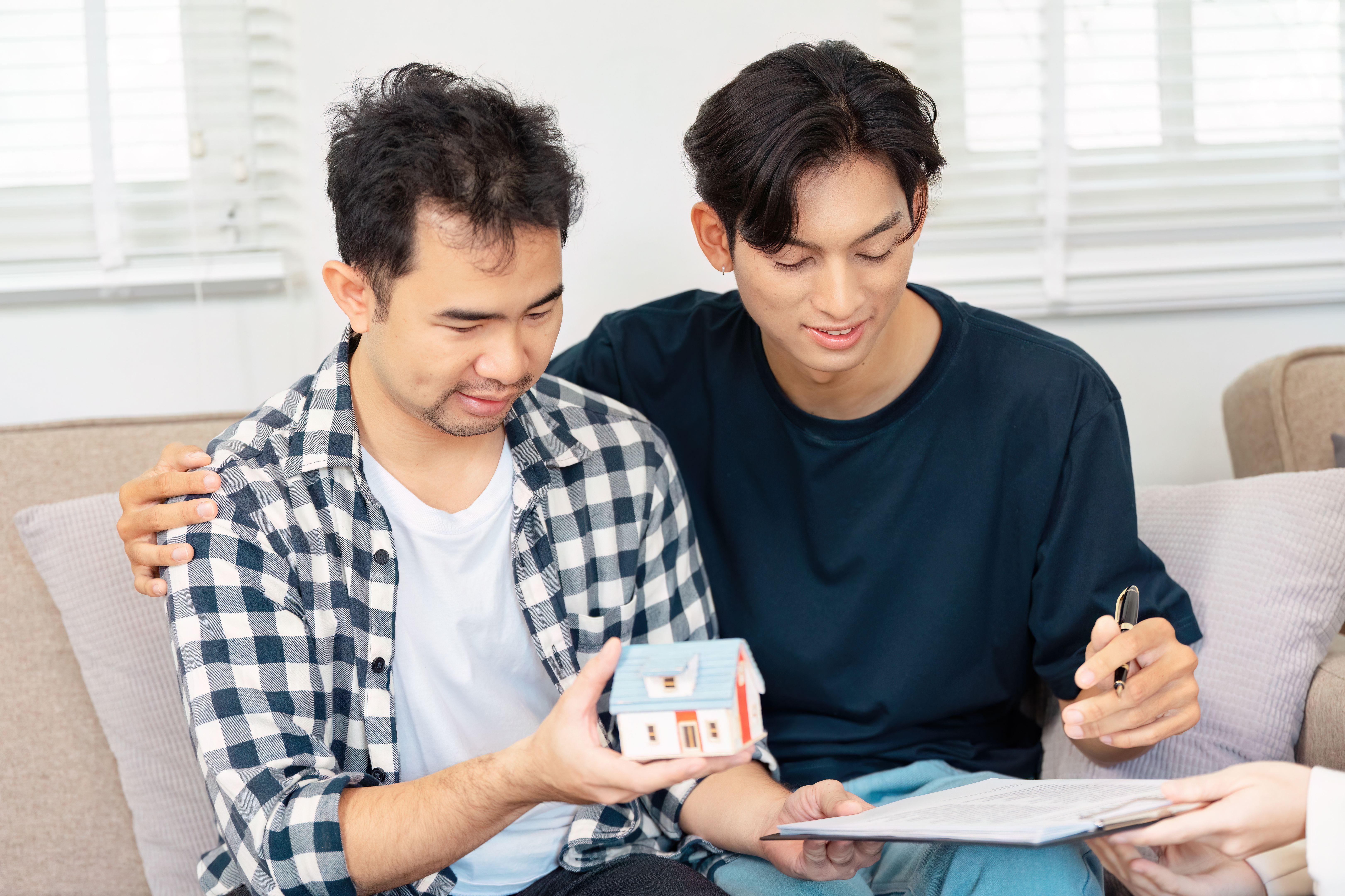 Diverse LGBTQ couple reviewing home purchase documents with model house. Diverse LGBTQ couple reviewing home purchase documents with model house.