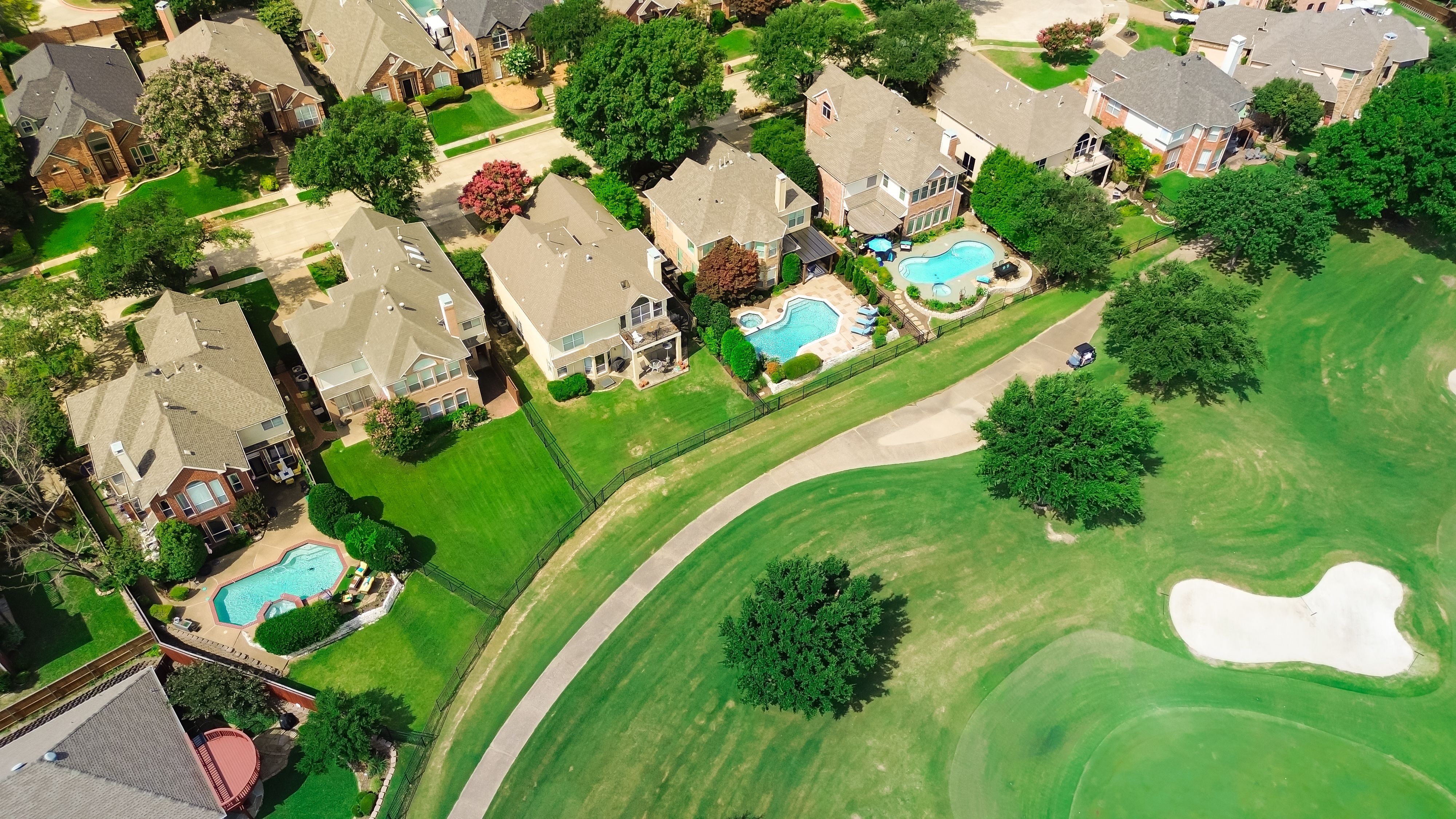 Upscale two-story suburban houses with swimming pools in golf course community in Plano, Texas, grassy country club with sand trap bunkers depression, club cars and golfers playing, aerial