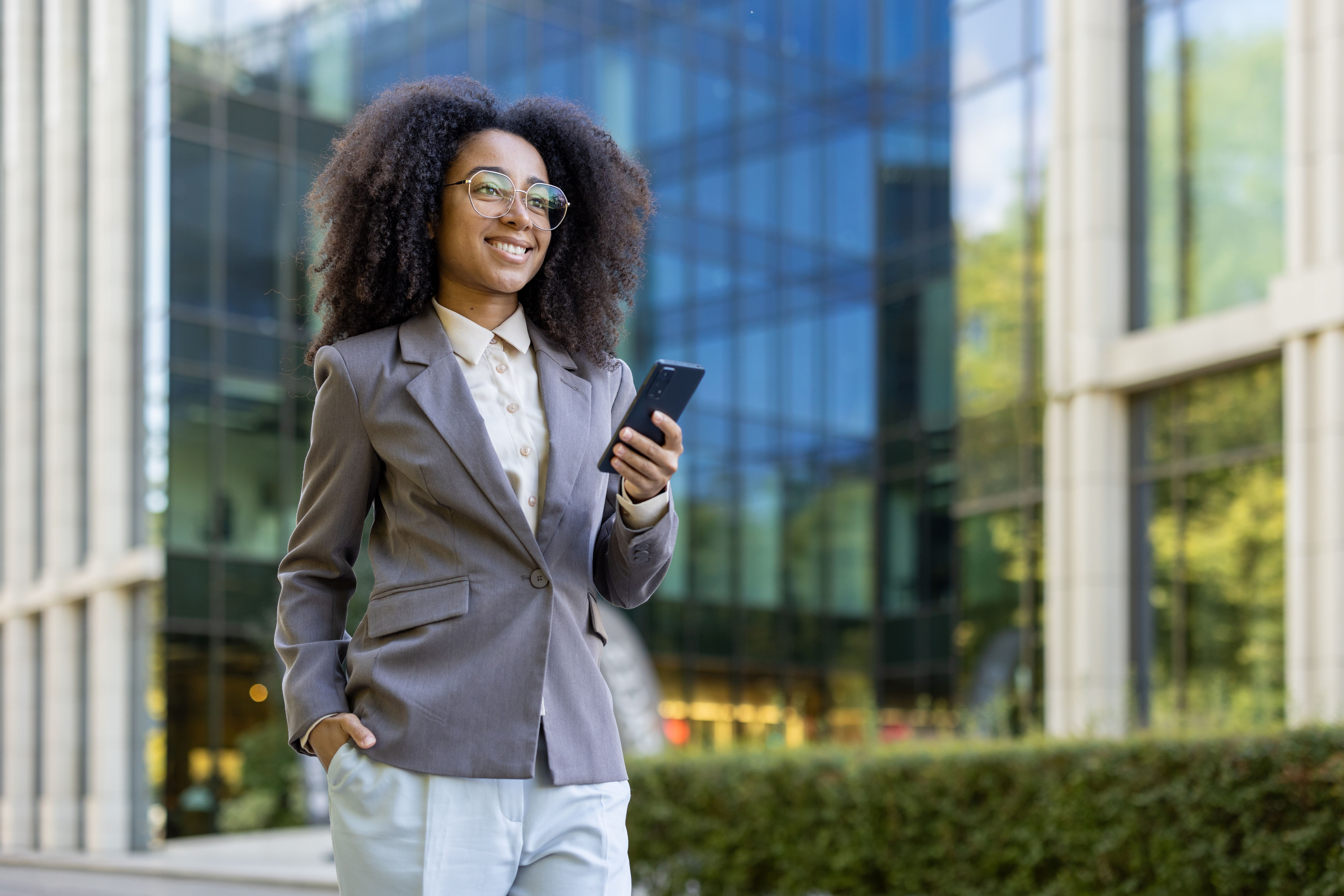 Confident African American businesswoman walking outside modern office building holding smartphone with positive energy