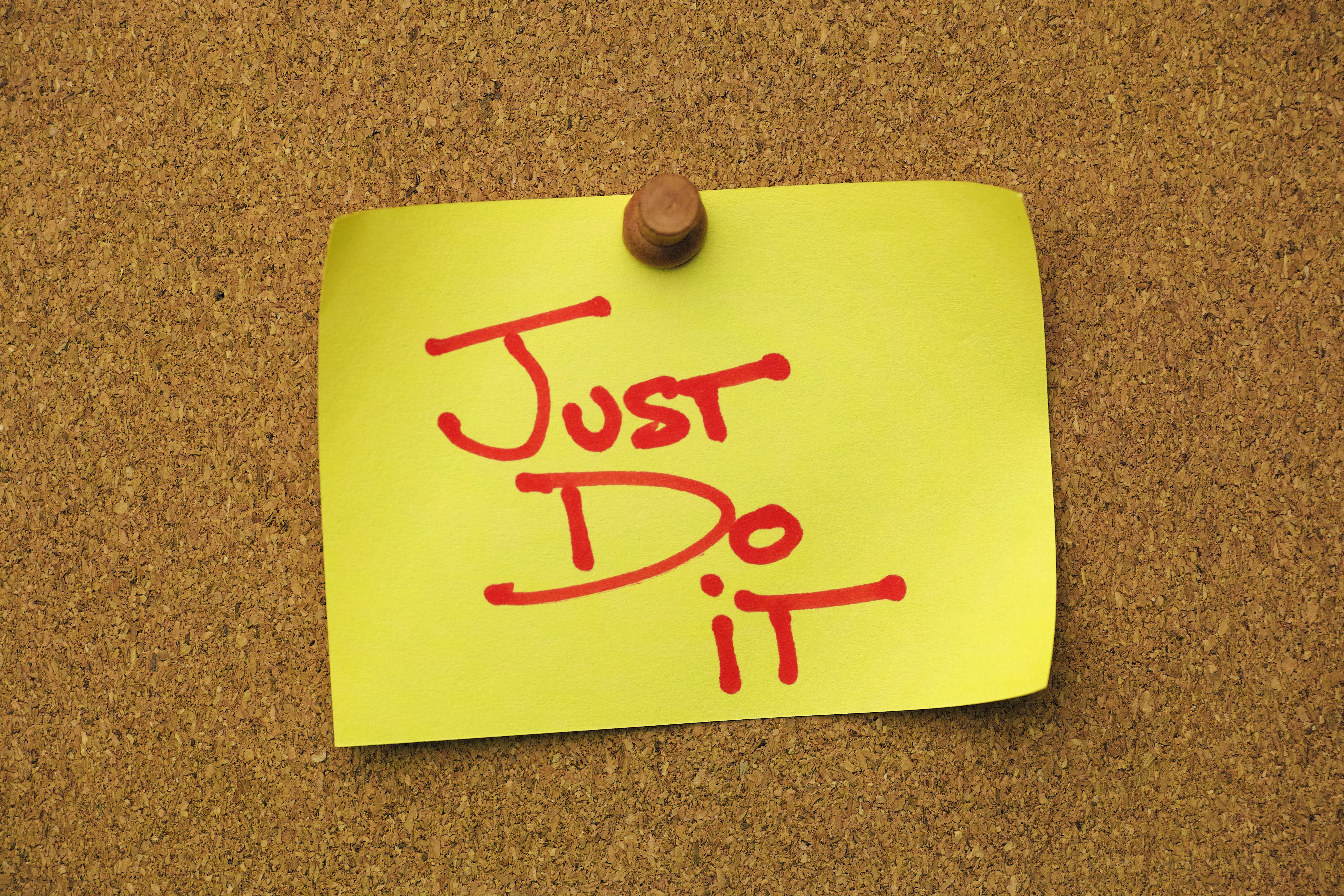 Yellow paper note pinned to a cork message board, with the short phrase 'JUST DO IT' written on it. Yellow paper note pinned to a cork message board, with the short phrase 'JUST DO IT' written on it.