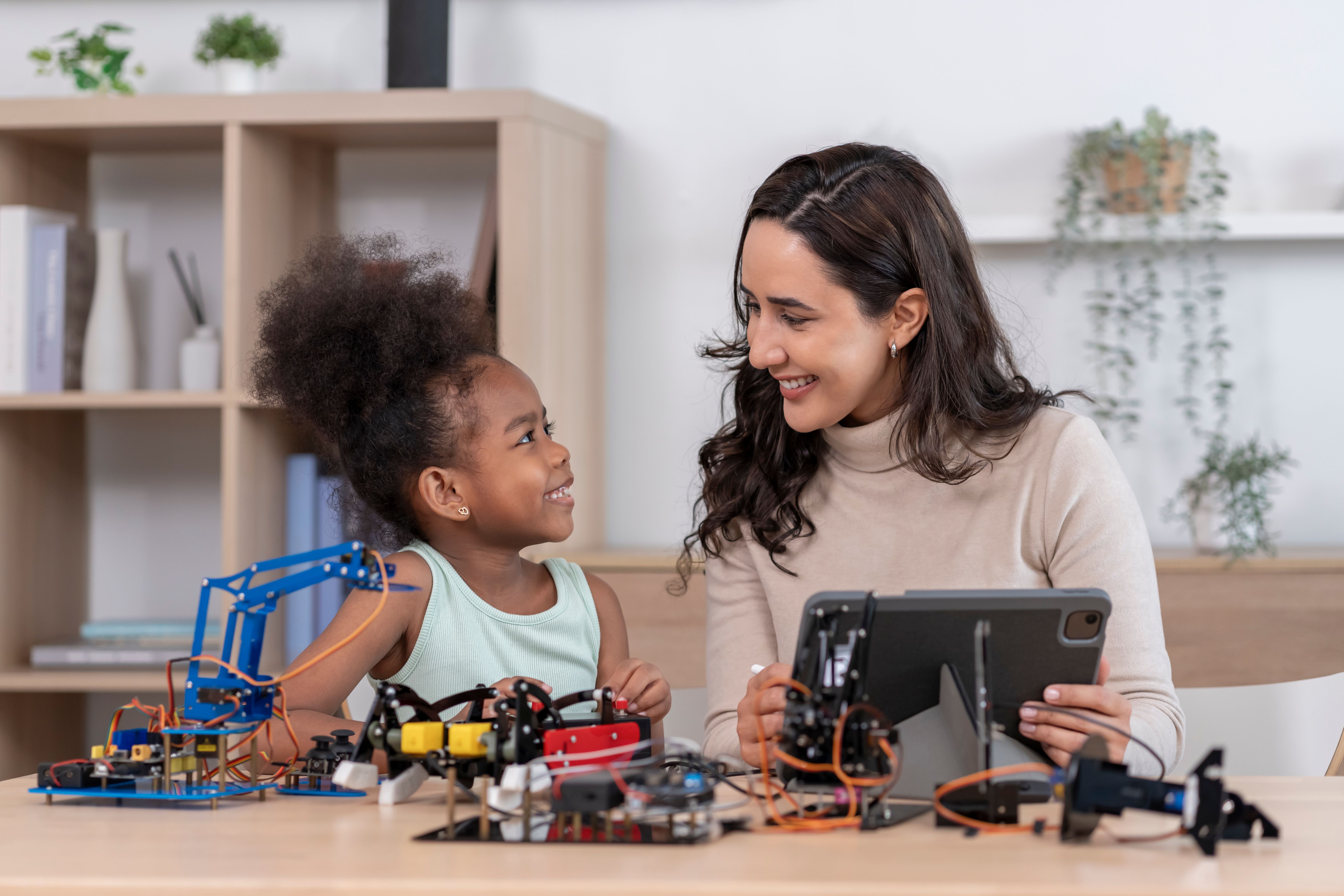 A caucasian female teacher holding digital tablet help african elementary school age girl working on robot circuit on table,STEM learning