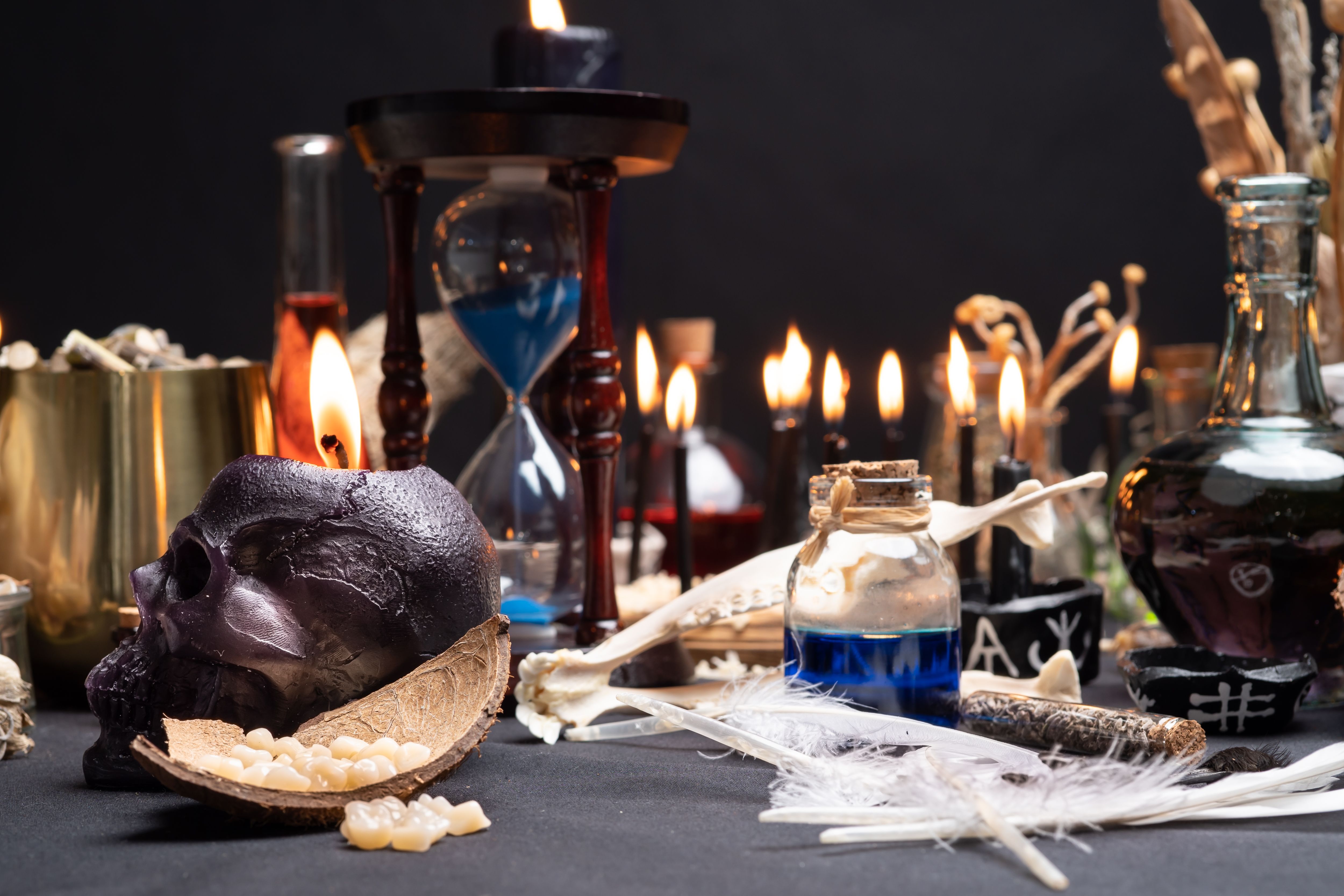 Witch table with witch craft magic items. Selective focus at front. Still life with black magic skull candle, ritual human teeth and bird feathers, potion vials and sand clock or hourglass. Halloween.