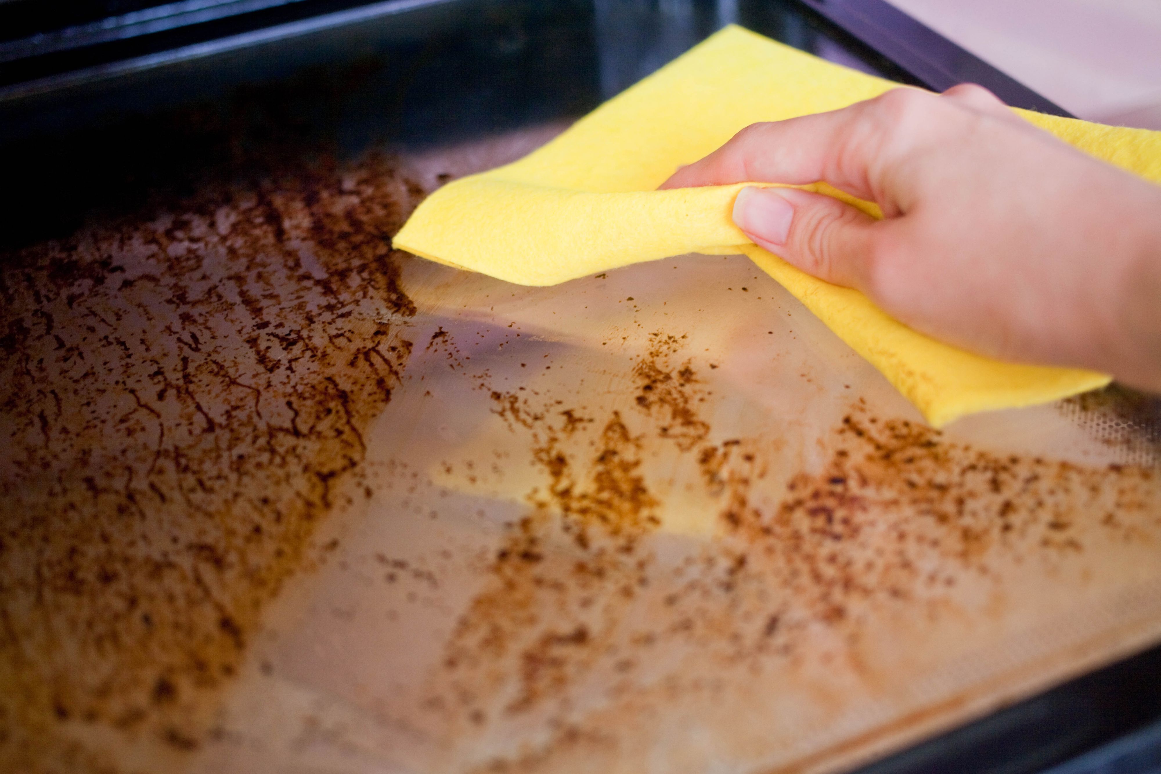 A female hand is holding a yellow rag and washing the dirty door of the oven. A female hand is holding a yellow rag and washing the dirty door of the oven.