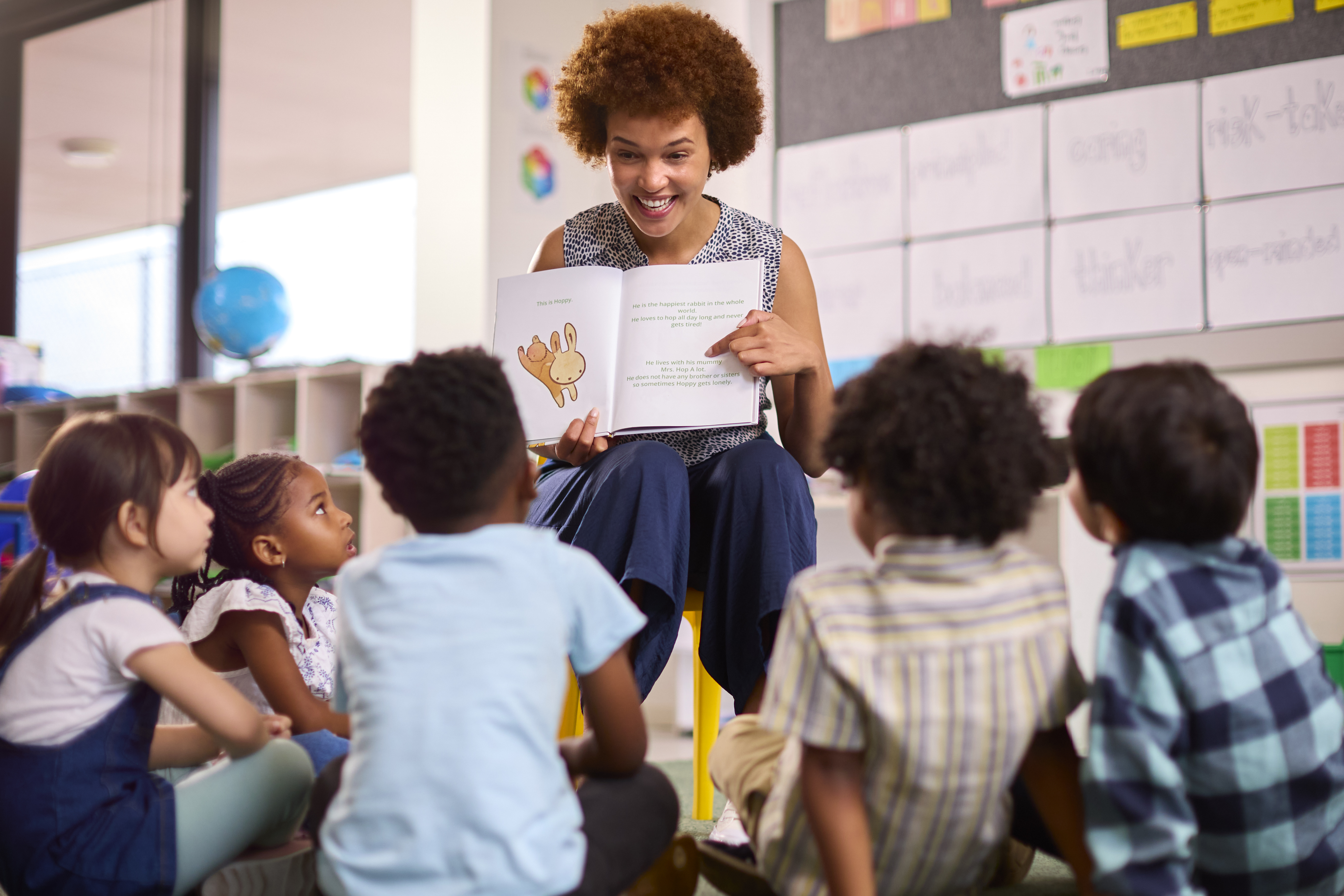 Female Teacher Reads To Multi-Cultural Elementary School Pupils Sitting On Floor In Class At School