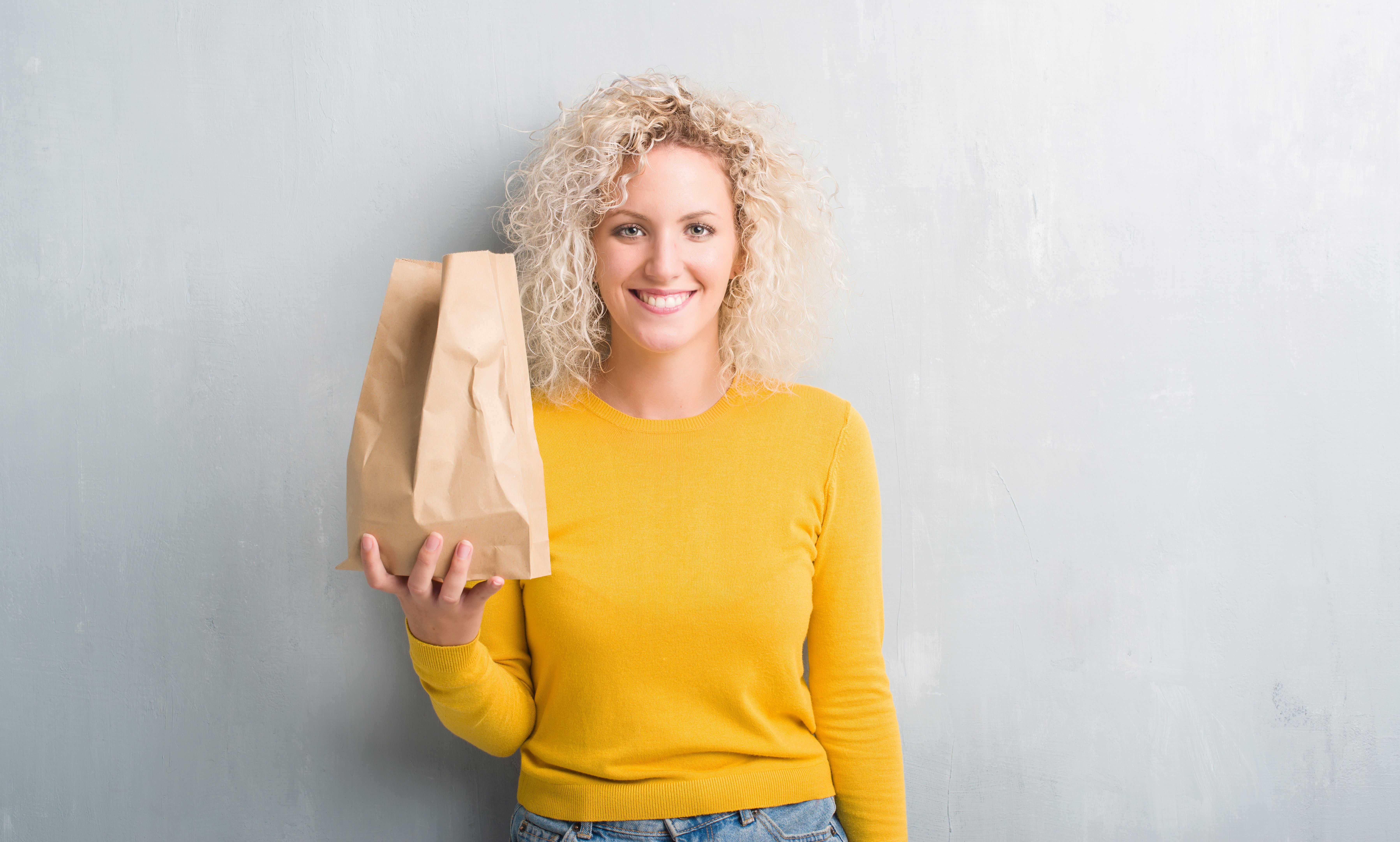 Young blonde woman over grunge grey background holding lunch paper bag with a happy face standing and smiling with a confident smile showing teeth Young blonde woman over grunge grey background holding lunch paper bag with a happy face standing and smiling with a confident smile showing teeth