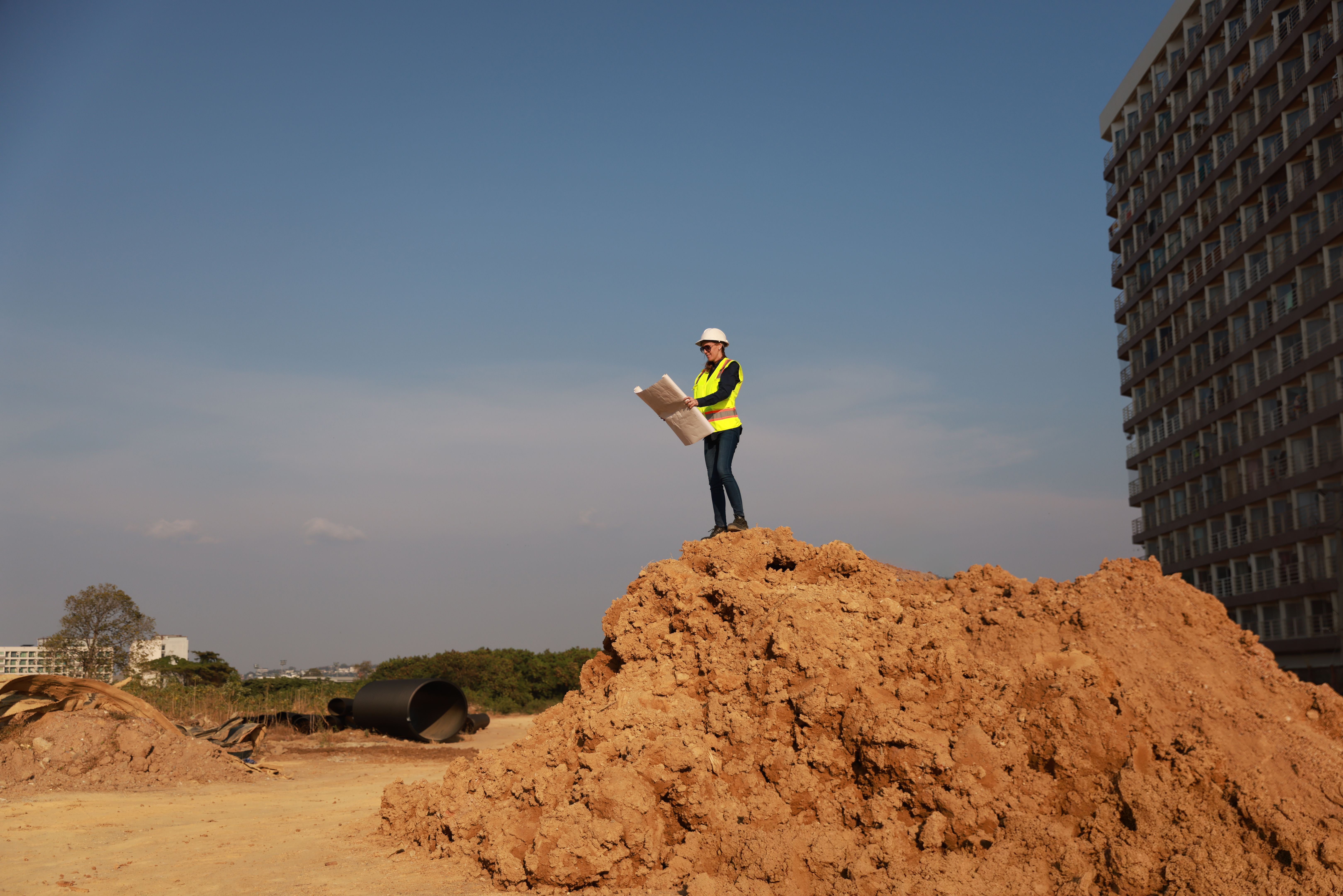 Low-angle view of a senior female  construction engineer, wearing full protective gear, examining a large blueprint map standing at the pile of dirt against the high resident building in the background