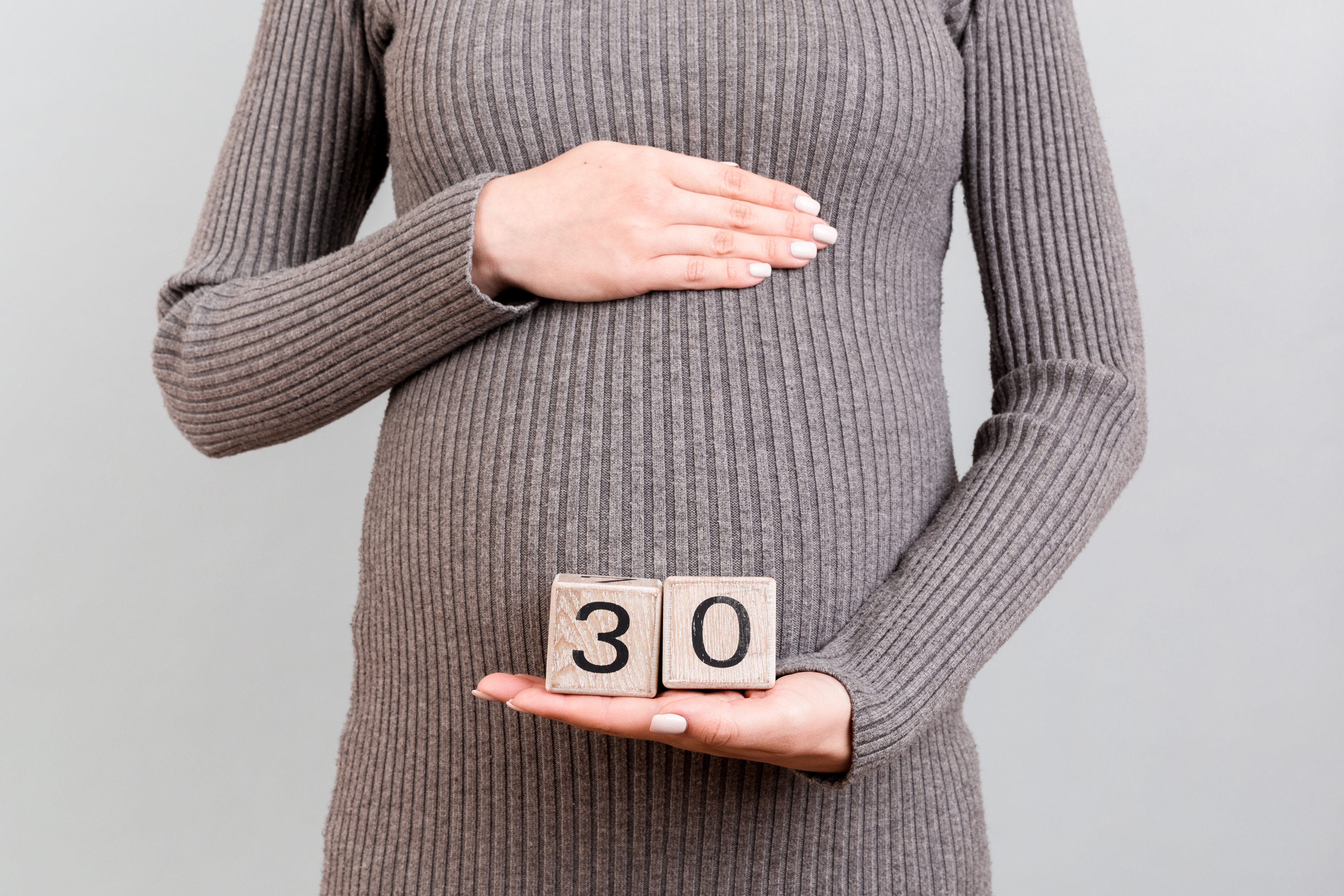 Close up of pregnant woman in gray dress holding cubes with numbers of pregnancy weeks. Thirty weeks of pregnancy period at gray background. Copy space