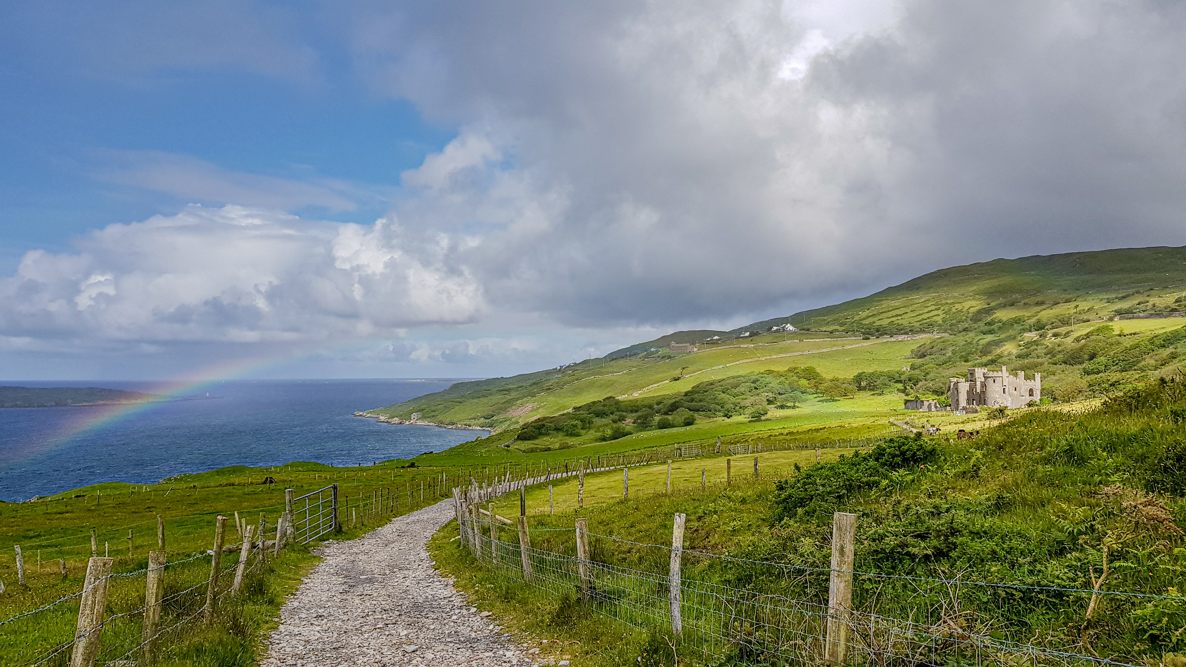Stone pathway with wooden pole fence and barbed wire between the Irish countryside and the sunlit Clifden Castle in the background