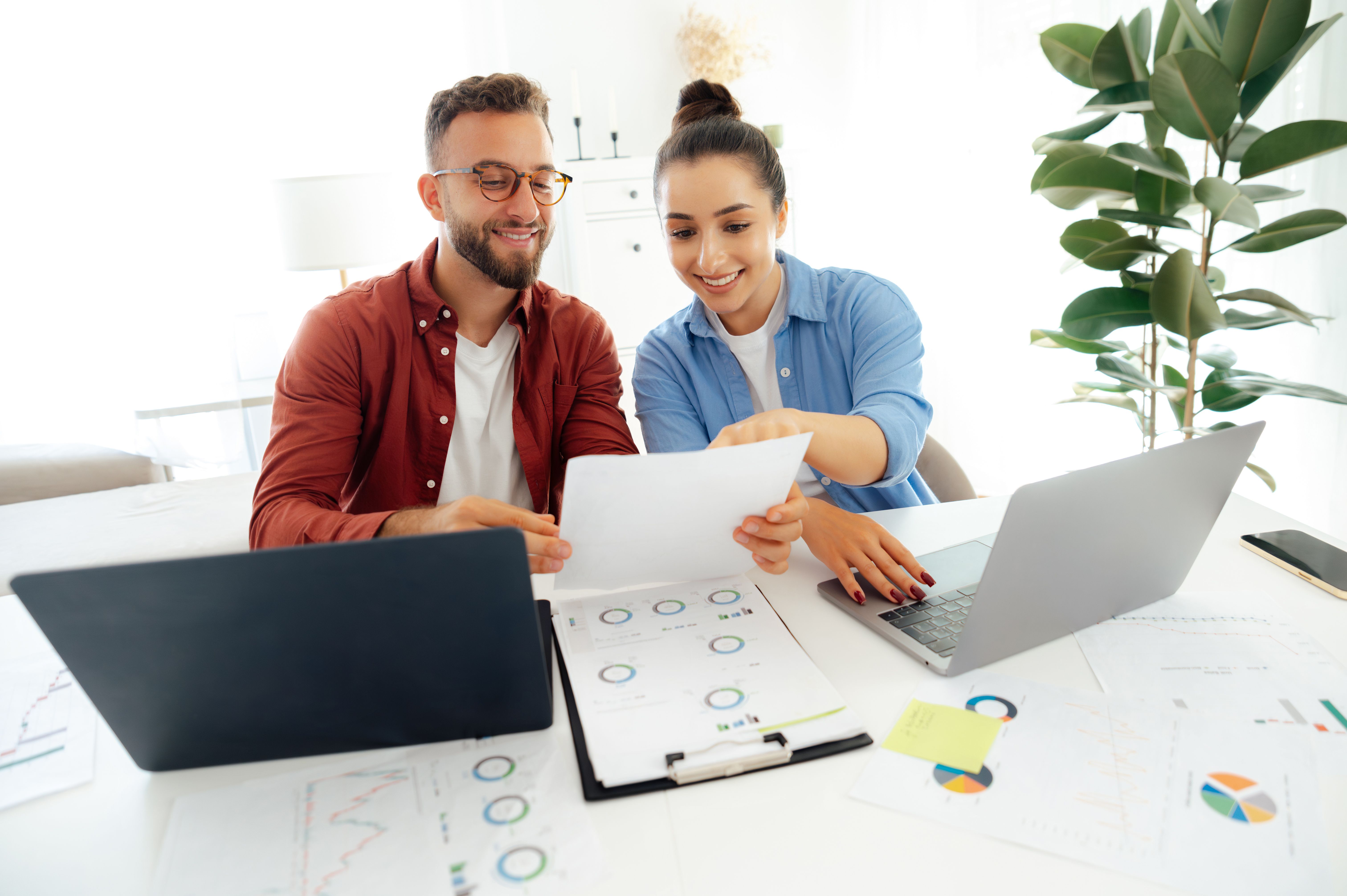 Team work collaboration. Two positive colleagues sit at their desks in an office, both working on laptops and smiling, they analyzing documents and discussing work-related matters, smile