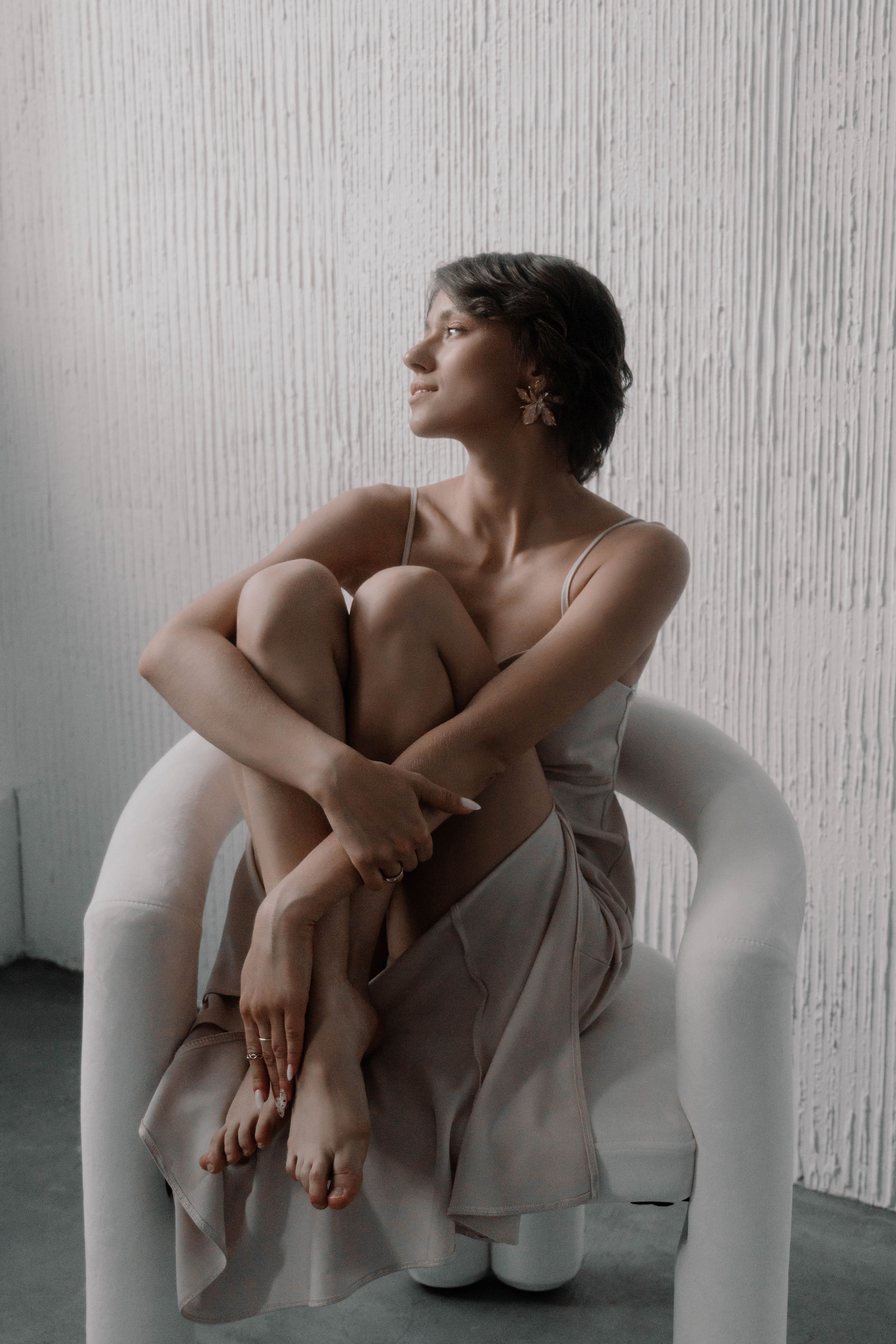 woman with short hair sits gracefully in a white chair, legs crossed, looking out a window with a serene expression. Soft light illuminates the simple yet stylish room