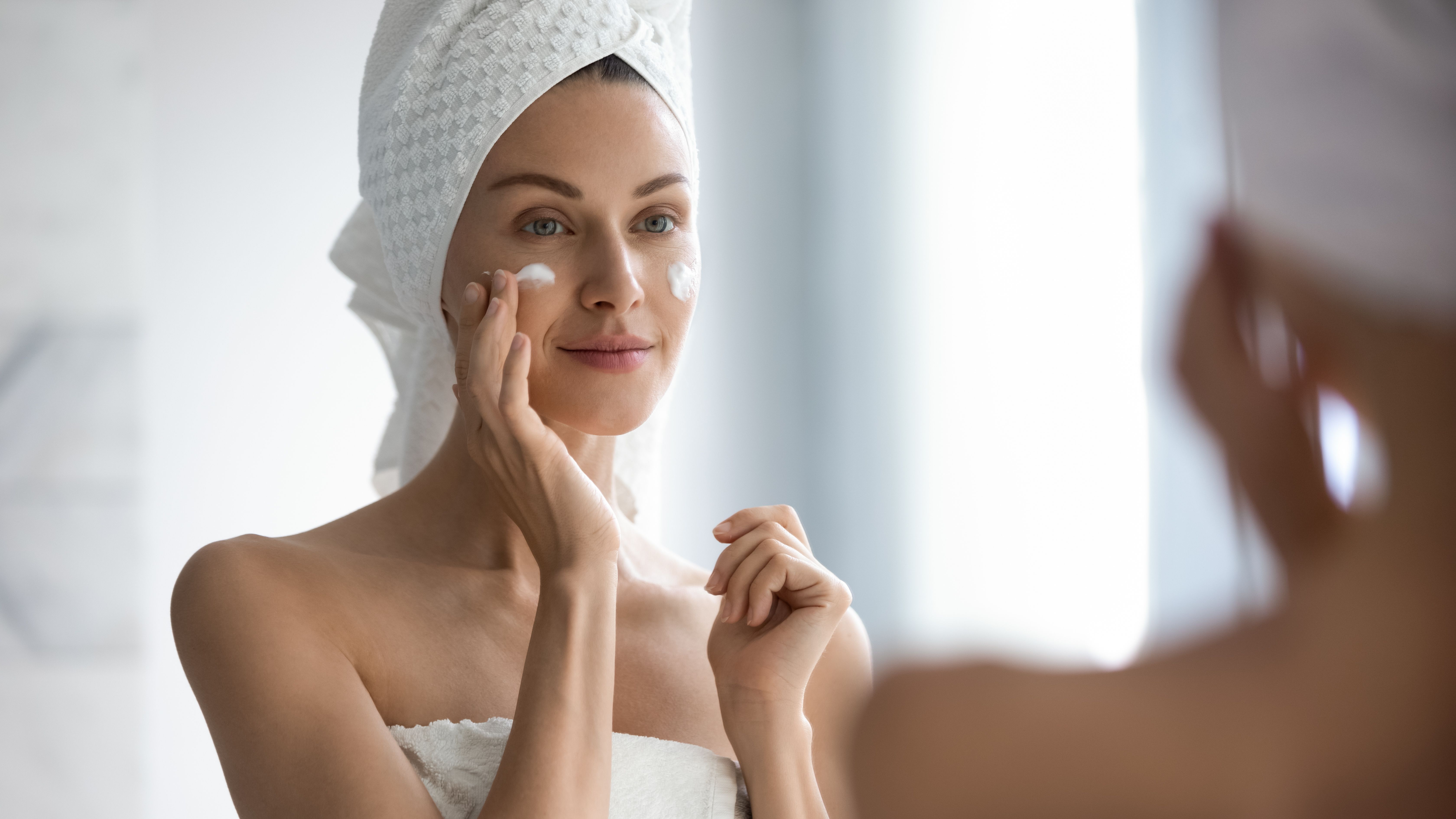 Attractive young adult woman applying facial cream looking in mirror Attractive young adult woman applying facial cream looking in mirror