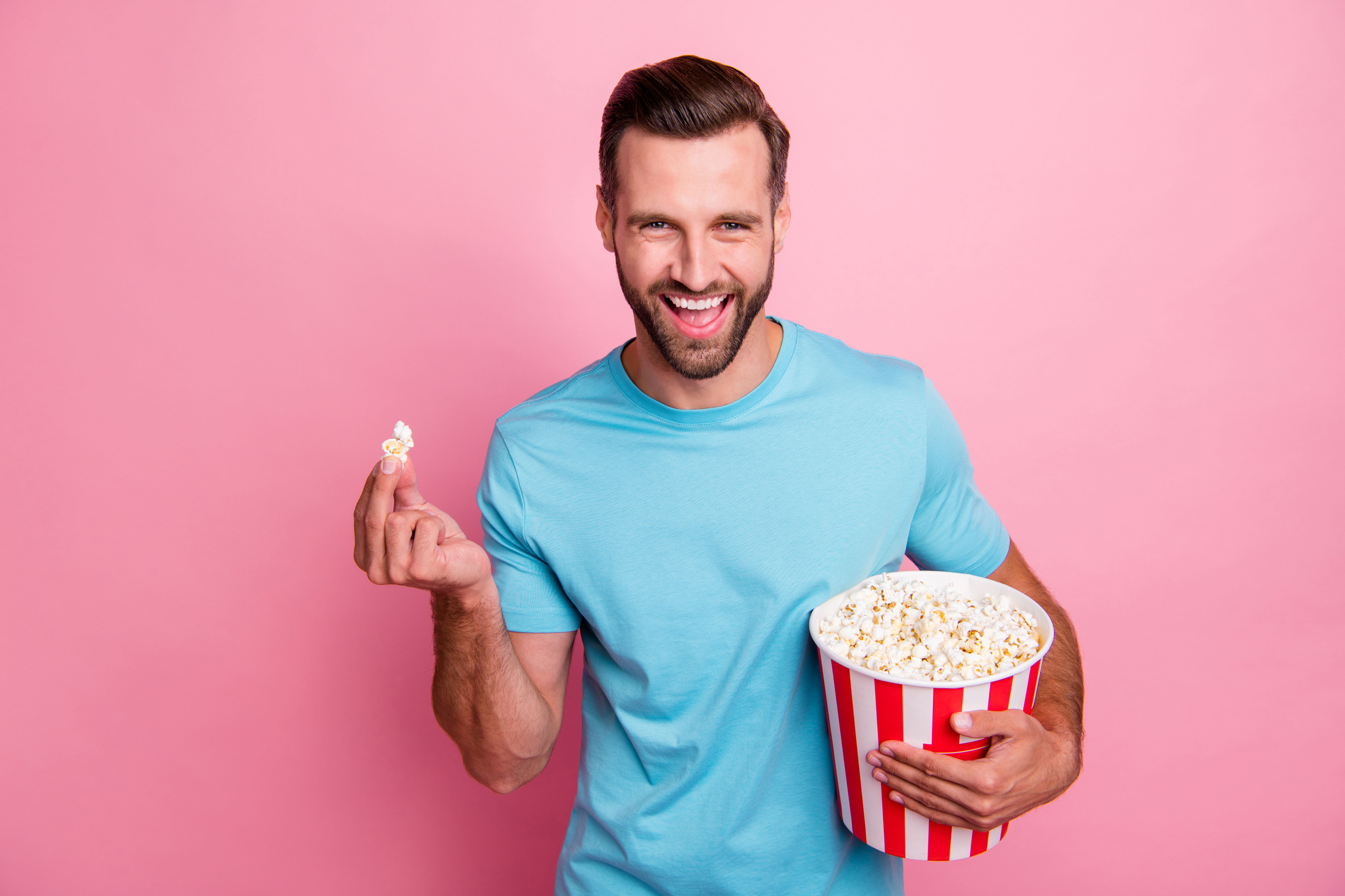 Photo of cheerful positive man watching tv series holding bucket of popcorn tasting smiling toothily comedy isolated in blue t-shirt over pastel color background