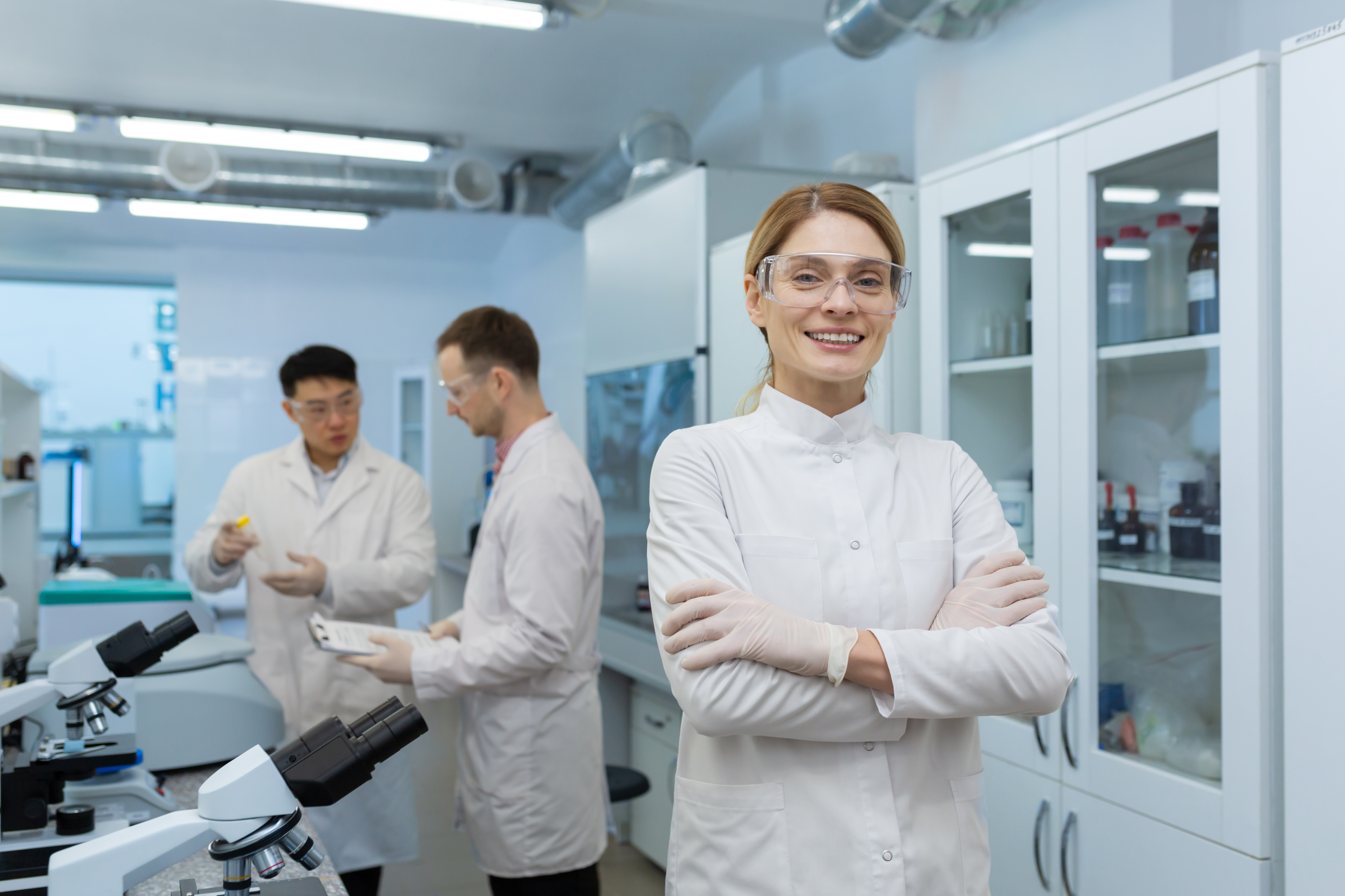 Portrait of a young woman scientist, chemist, doctor standing in the laboratory of the clinic, crossing her arms on her chest and smiling at the camera. In the background, male colleagues