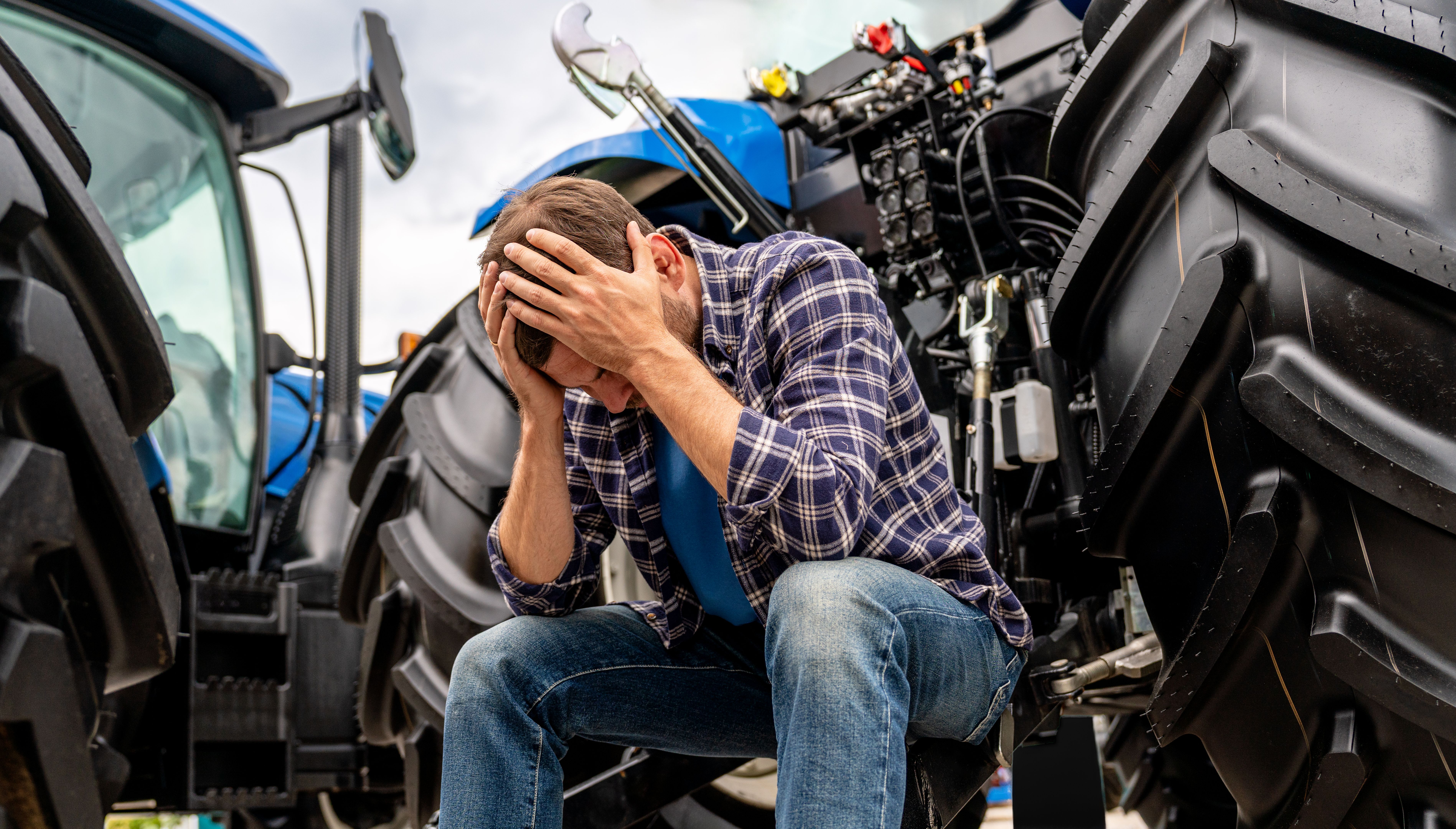 Male farmer with head in hands by tractor as symbol of ongoing farm crisis