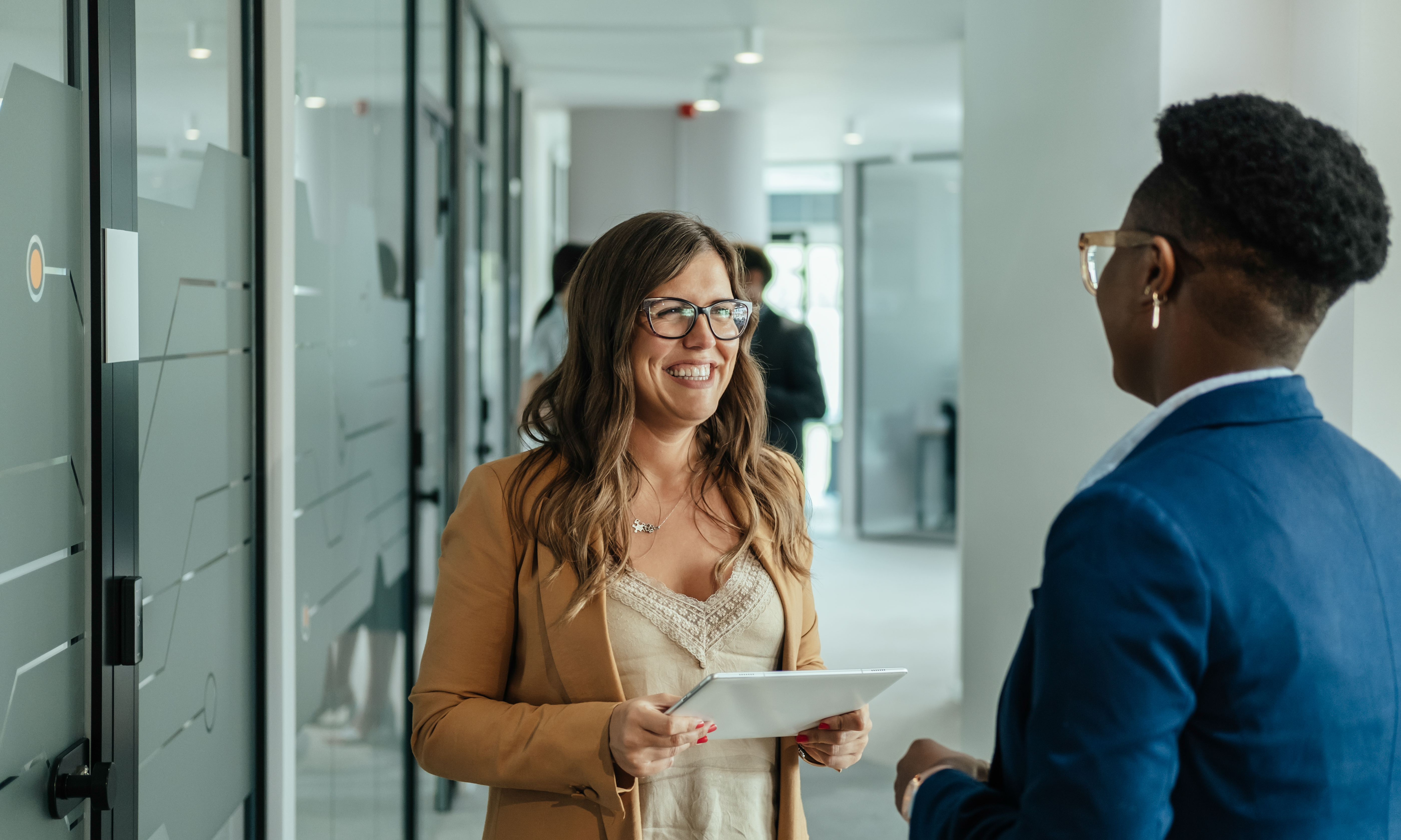 Happy Businesswoman Holding Digital Tablet and Talking with her Anonymous Female Colleague at Work