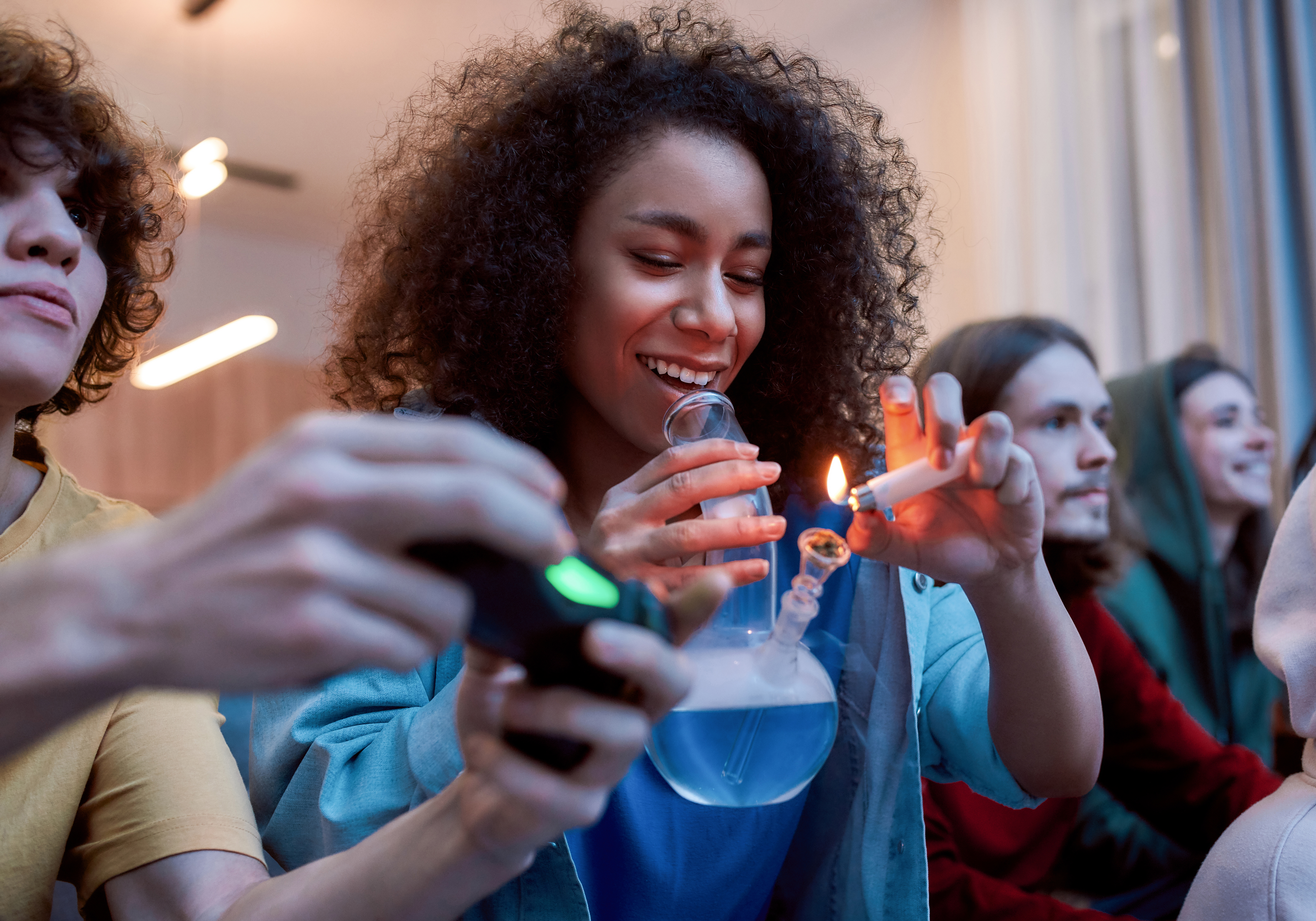 For great mood. Young afro american girl lighting marijuana in the glass bong, relaxing with friends on the sofa at home. Young people playing video games and smoking weed
