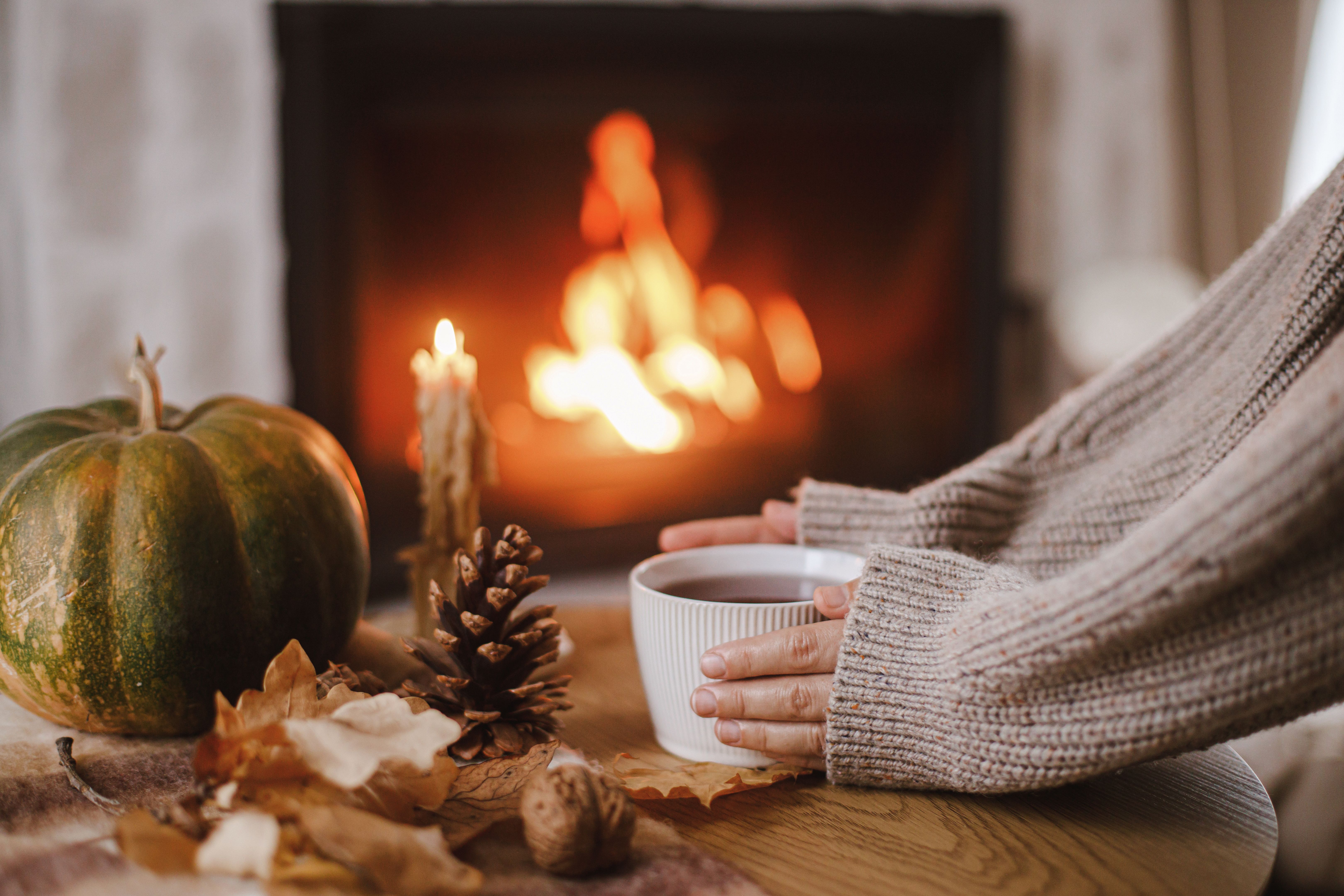 Hands in cozy sweater holding warm cup of tea with pumpkin and autumn leaves on table on background of burning fireplace. Autumn hygge. Thanksgiving and Halloween.