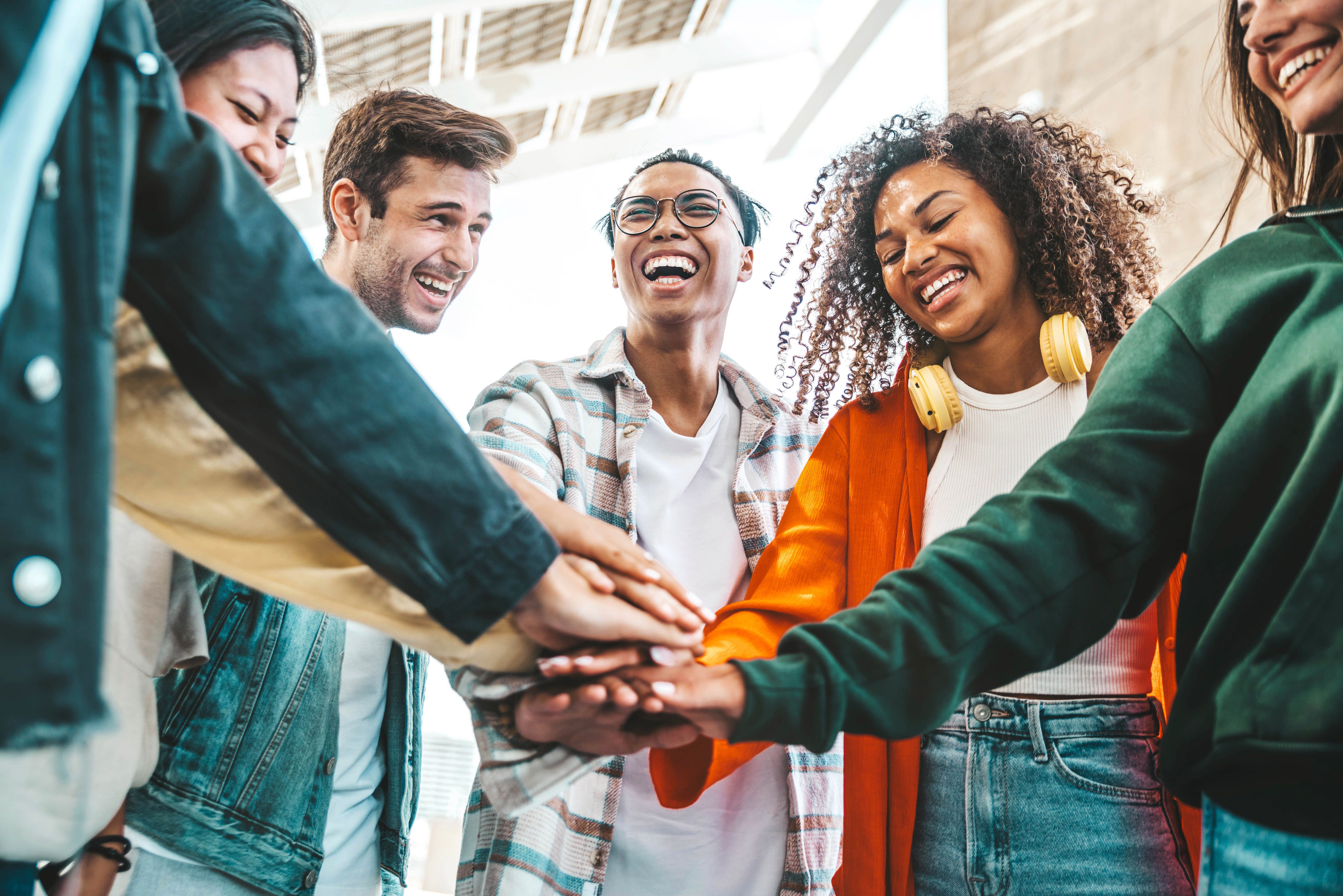 Multiracial group of young people stacking hands outdoors - Happy friends celebrating success on city street - Youth community concept with guys and girls standing together supporting peace and love