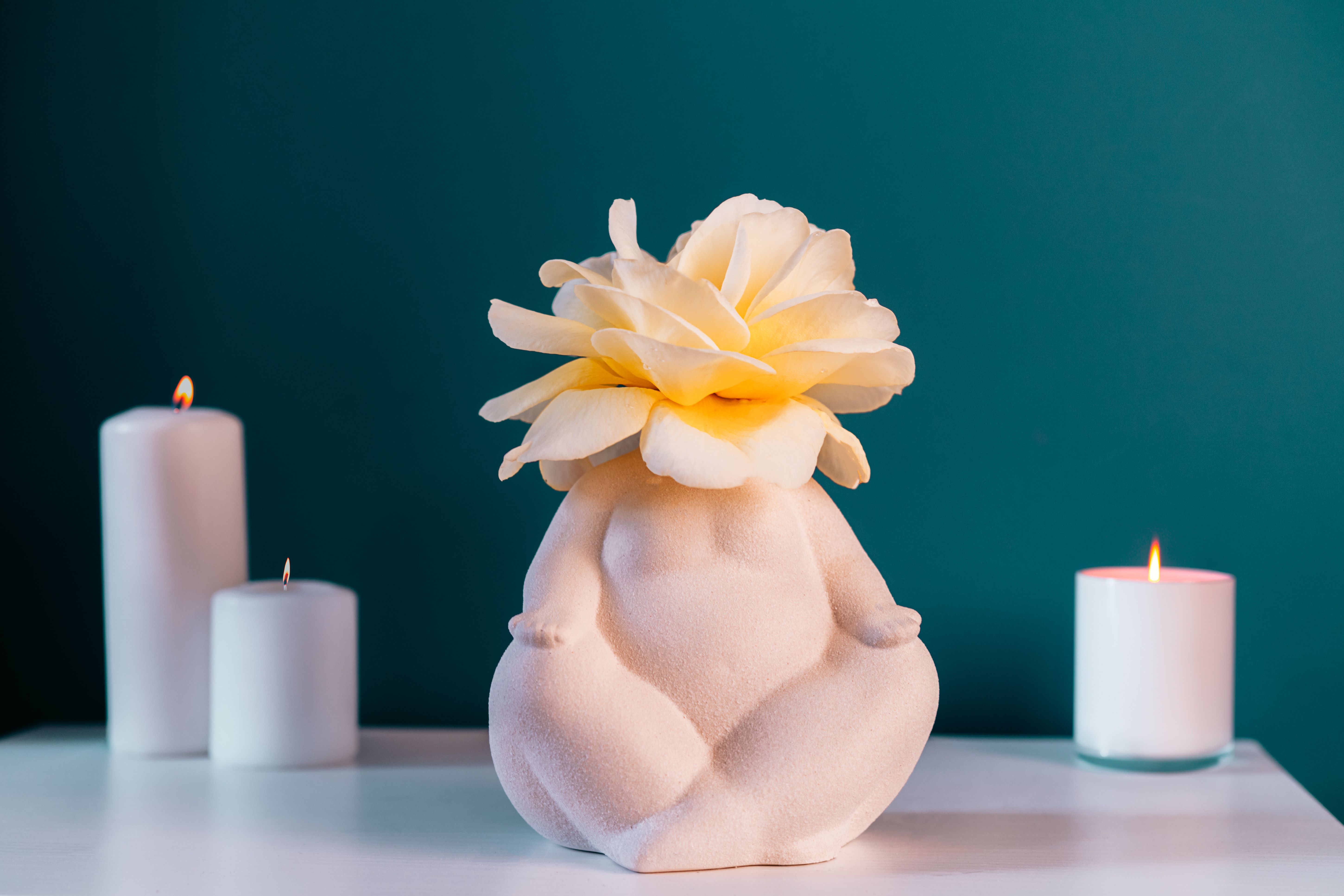 Close up white table with ceramic female body-shaped vase with rose flower, burning candles on turquoise blue wall background. Zen, cozy, meditation female bedroom decor. Self recovery place at home.