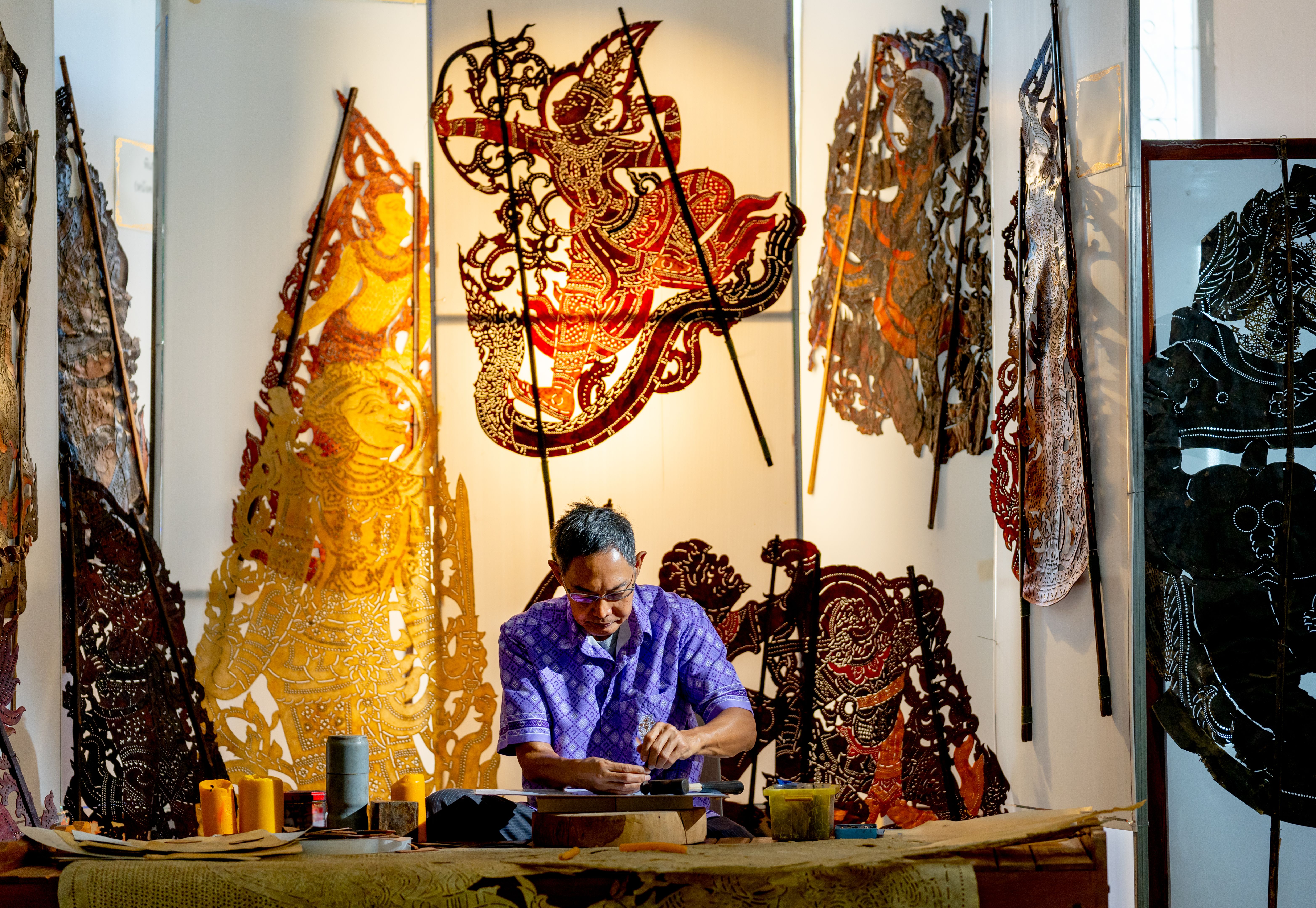 Front view Asian senior man wear glasses use hammer and chisel to create grand shadow play craft and he concentrate to work in area of workplace with several products are place around workplace area.