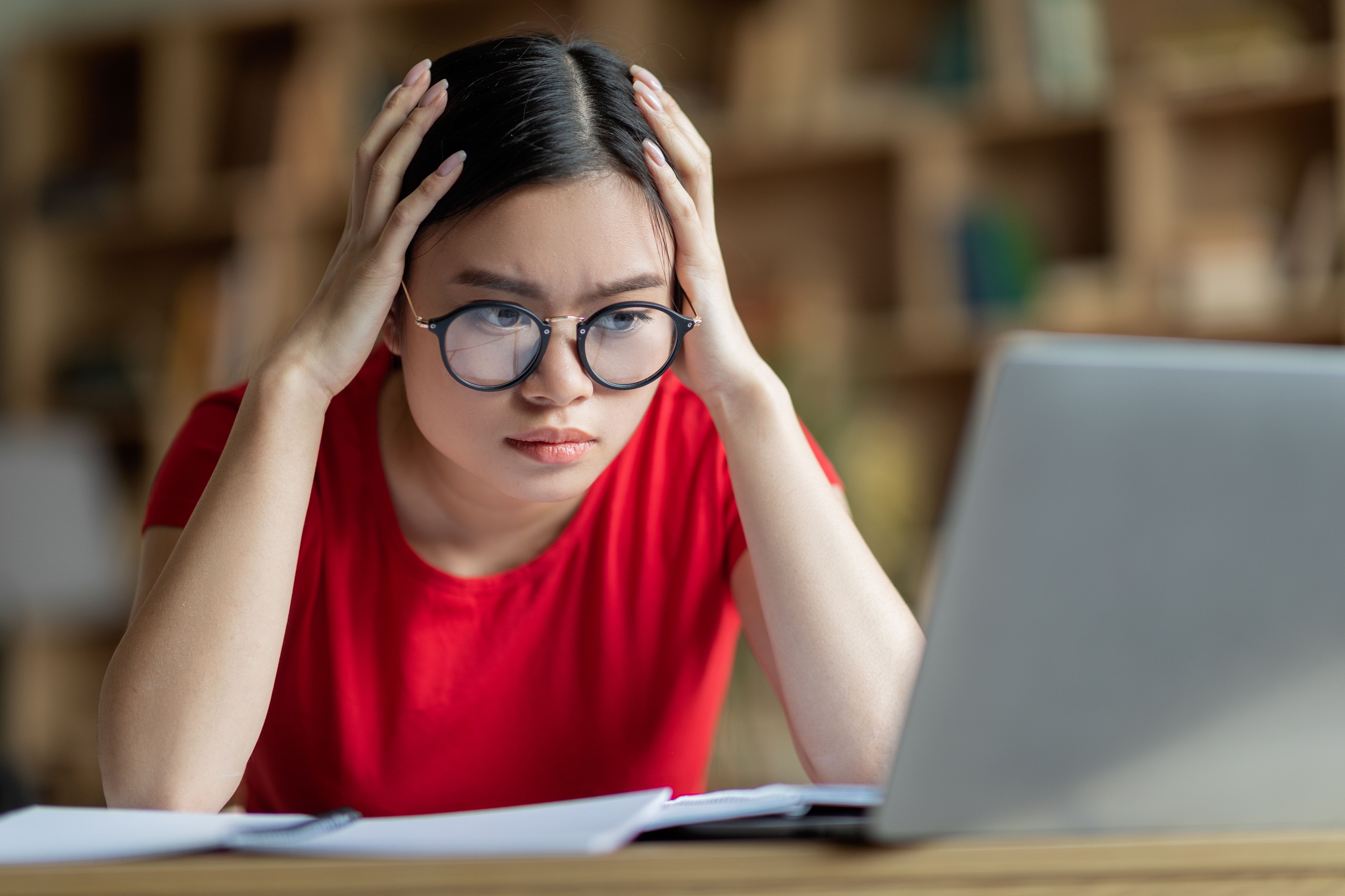 Concentrated busy young asian girl in glasses hold head with hands, look at computer in room interior