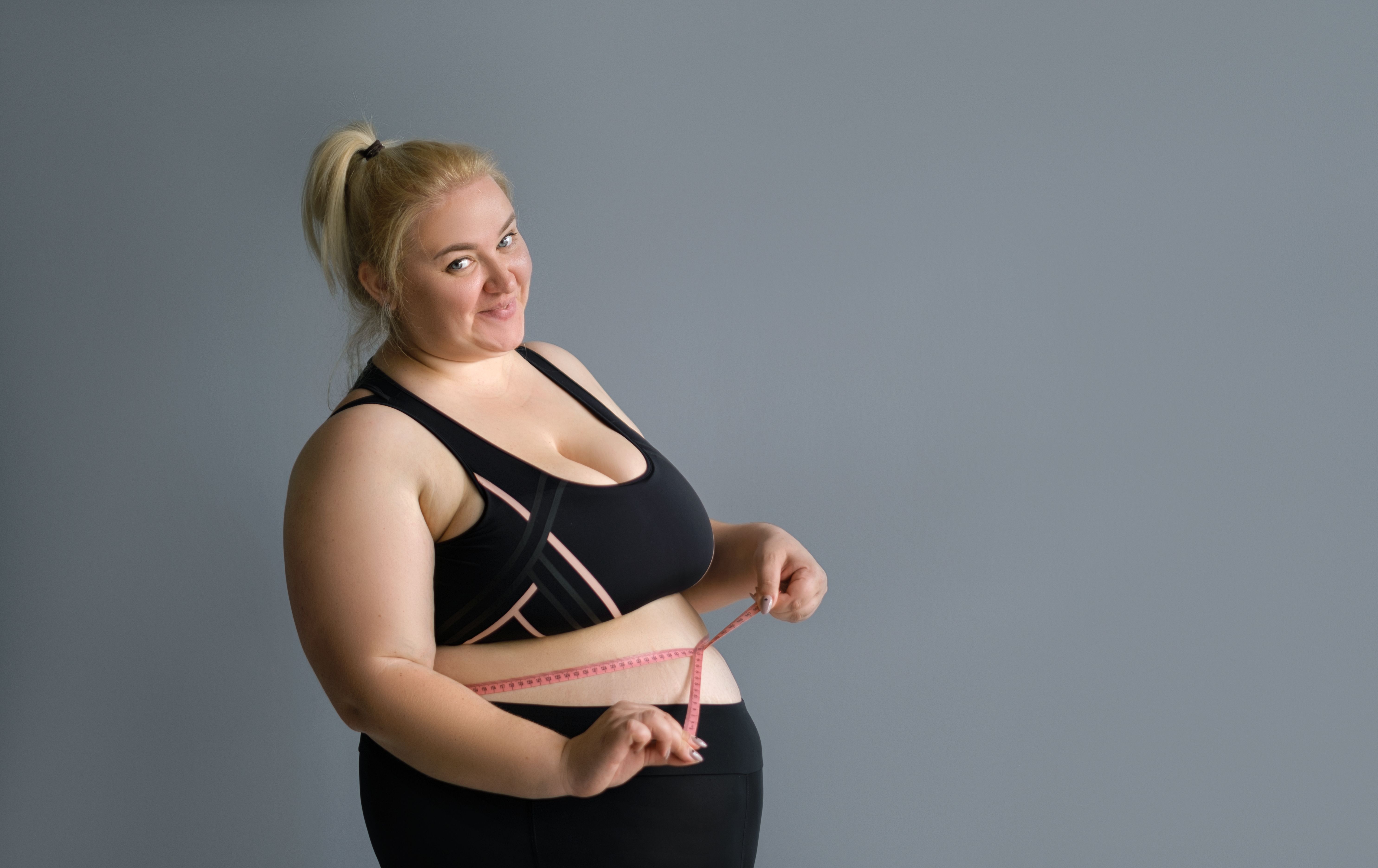 A fat big lady measures the waist measuring tape on a gray studio background. The woman is happy with the result of the diet and lost a couple of extra pounds. Copy space A fat big lady measures the waist measuring tape on a gray studio background. The woman is happy with the result of the diet and lost a couple of extra pounds. Copy space