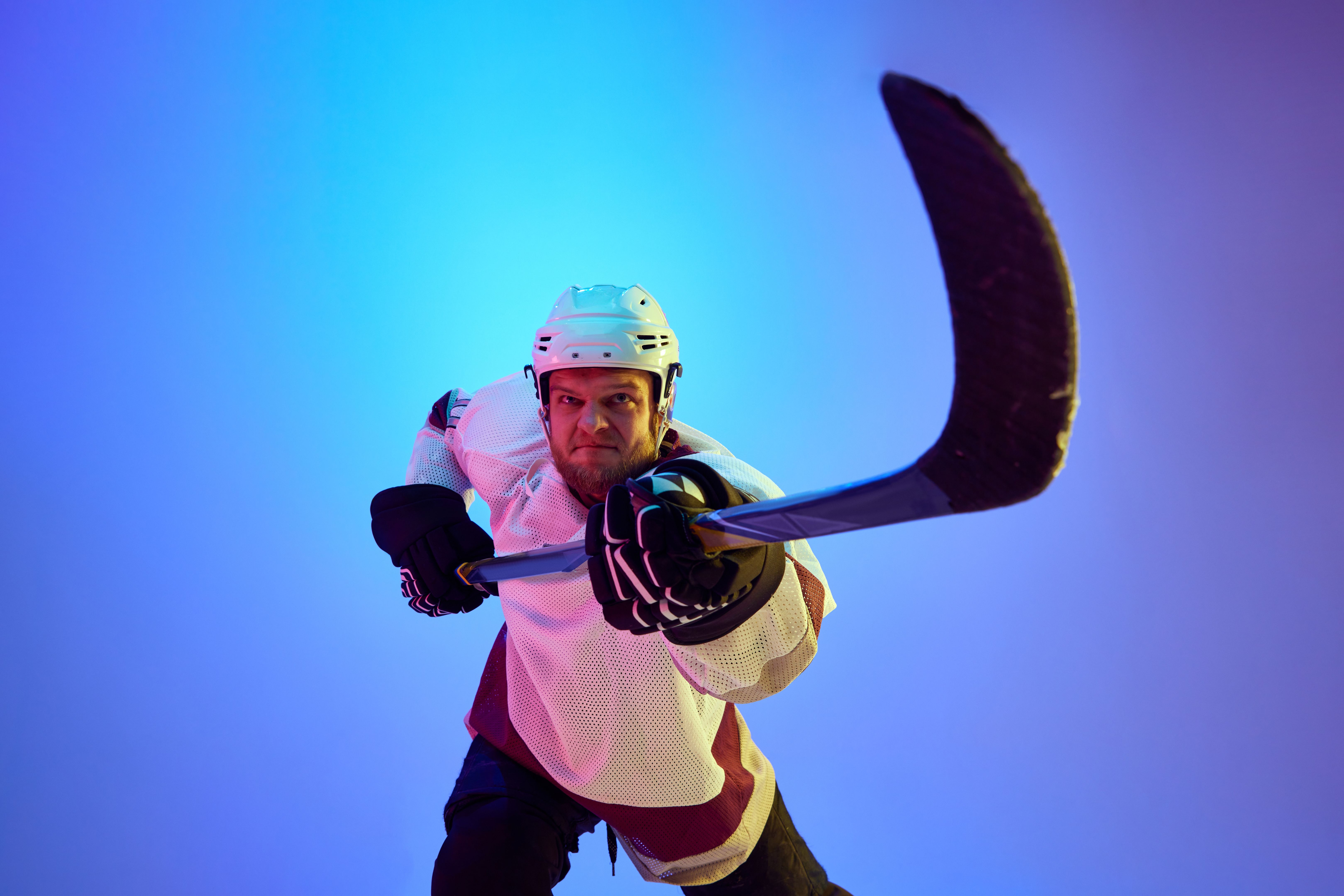Competitive, motivated man, hockey player posing with stick, in uniform and helmet against gradient blue background in neon light