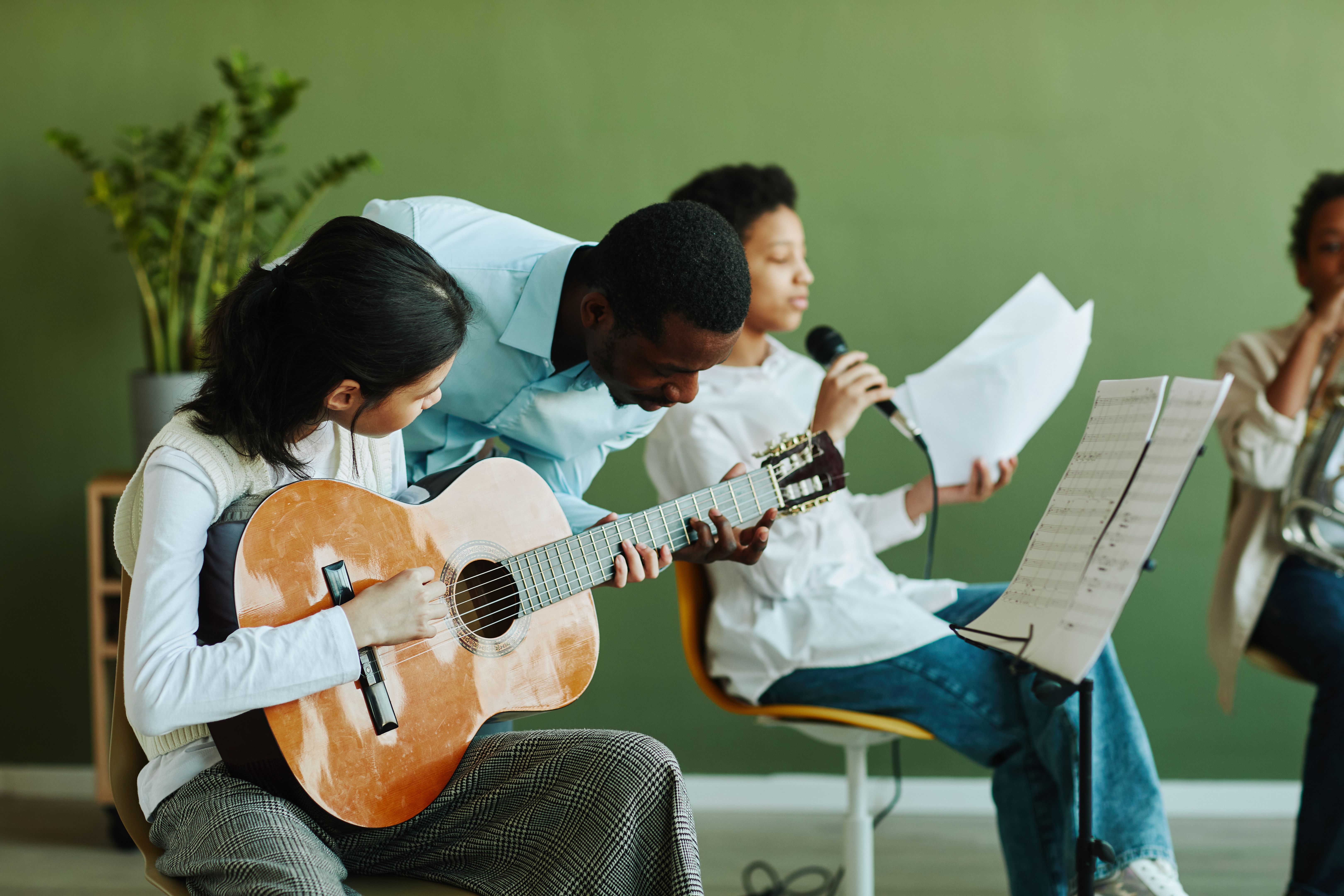 Young teacher of music bending over Asian schoolgirl with acoustic guitar