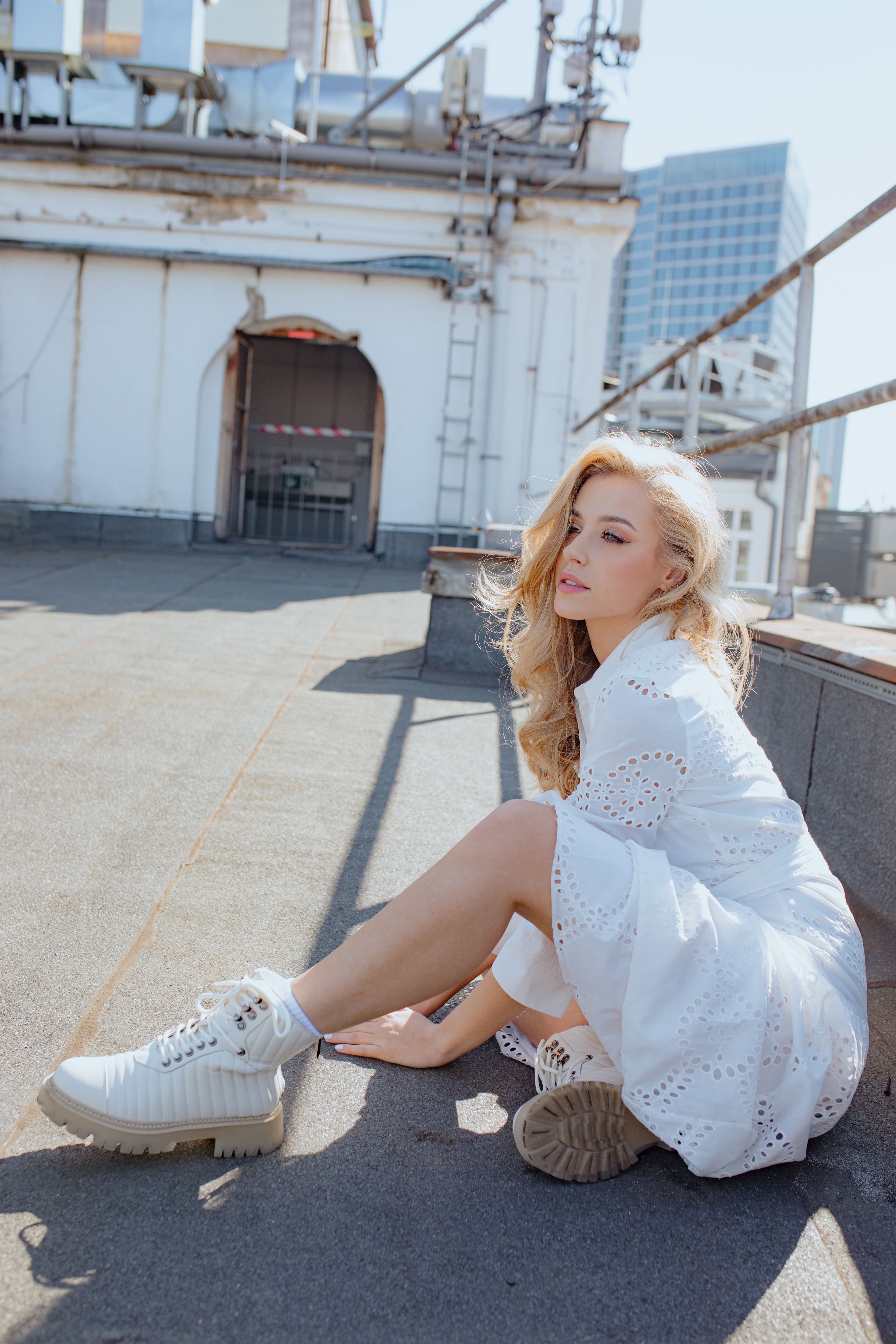 Young woman in white lace dress, laced boots sitting near fence on solar rooftop of city building and looking forward.